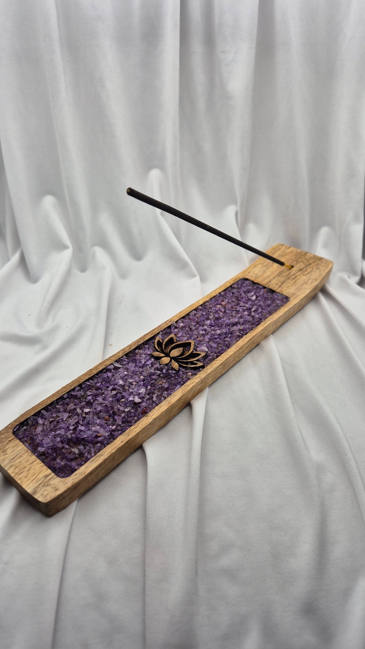 A rectangular wooden incense holder with a purple crystal insert and a single incense stick resting diagonally across it, set against a draped white fabric background.