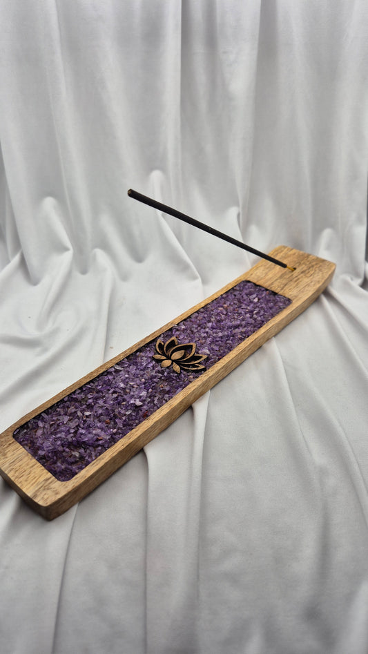 A rectangular wooden incense holder with a purple crystal insert and a single incense stick resting diagonally across it, set against a draped white fabric background.