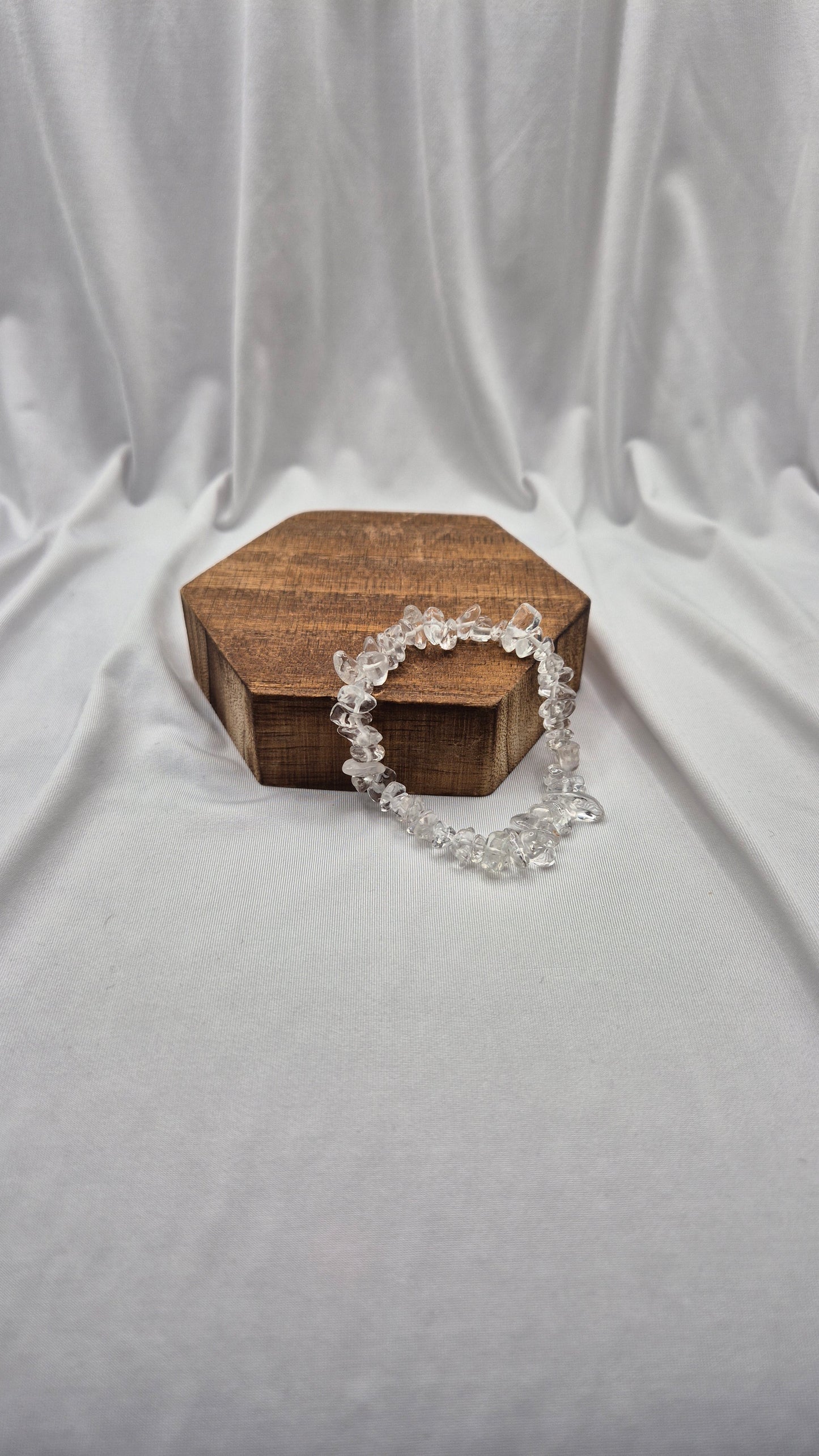A clear crystal beaded bracelet draped over a small hexagonal wooden box, set against a draped white fabric backdrop.