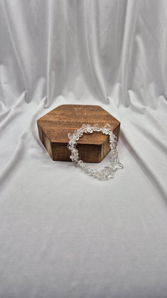 A clear crystal beaded bracelet draped over a small hexagonal wooden box, set against a draped white fabric backdrop.