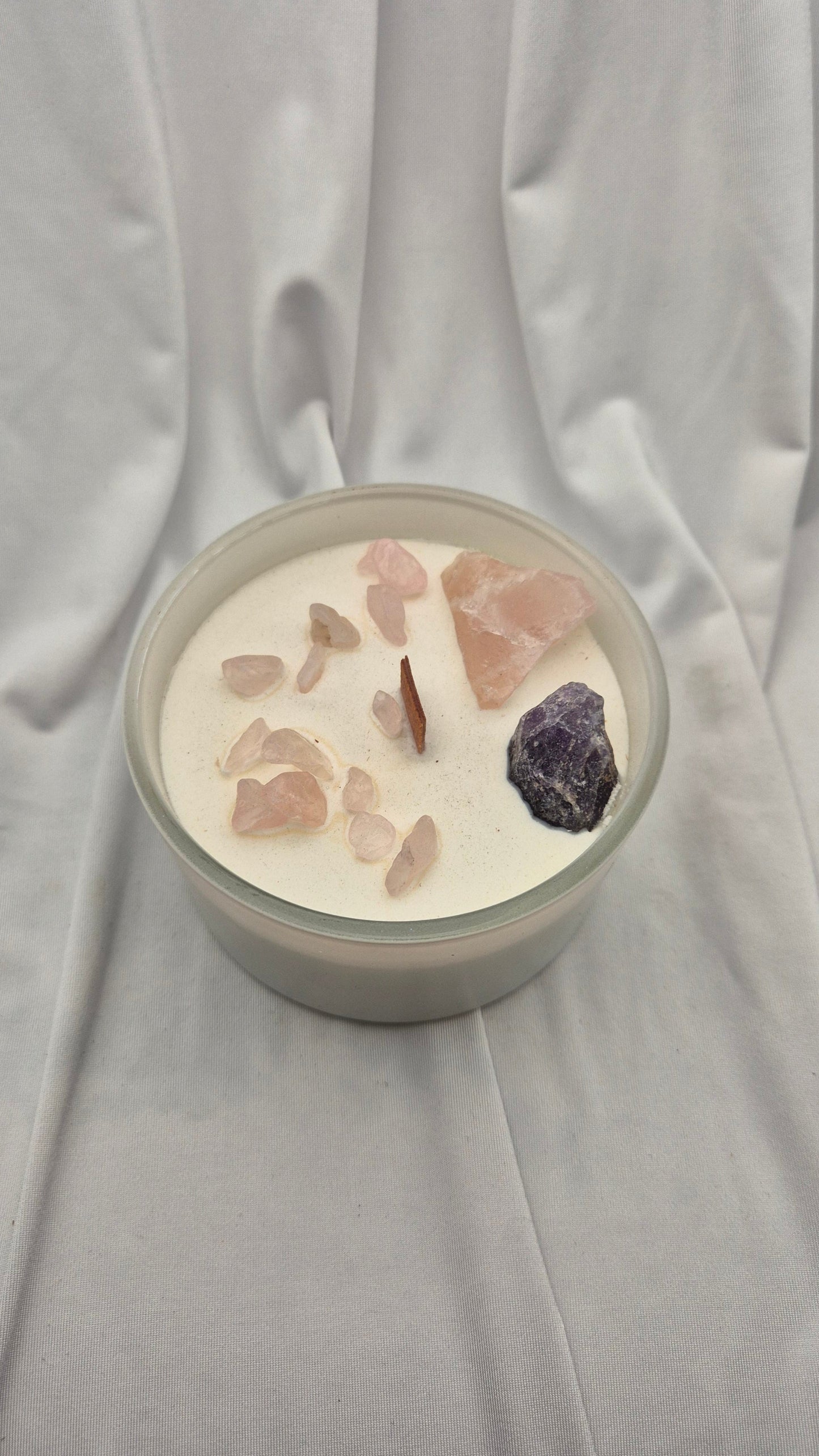 A glass jar candle filled with white wax, decorated with pink rose quartz crystals and a dark purple amethyst crystal, set against a draped white fabric background.