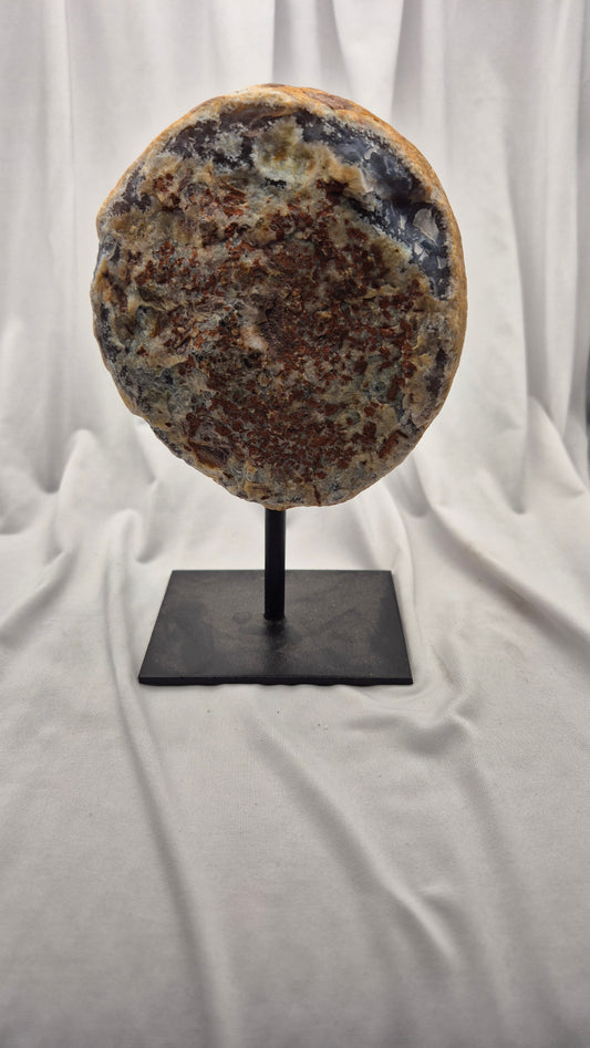 A large circular polished stone or geode slice displayed upright on a black metal stand against a draped white fabric backdrop.