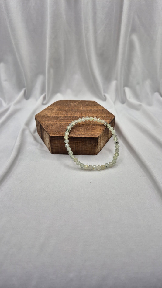 A beaded stretch bracelet made of small, pale greenish-white round beads, displayed on a small hexagonal wooden box against a draped white fabric backdrop.