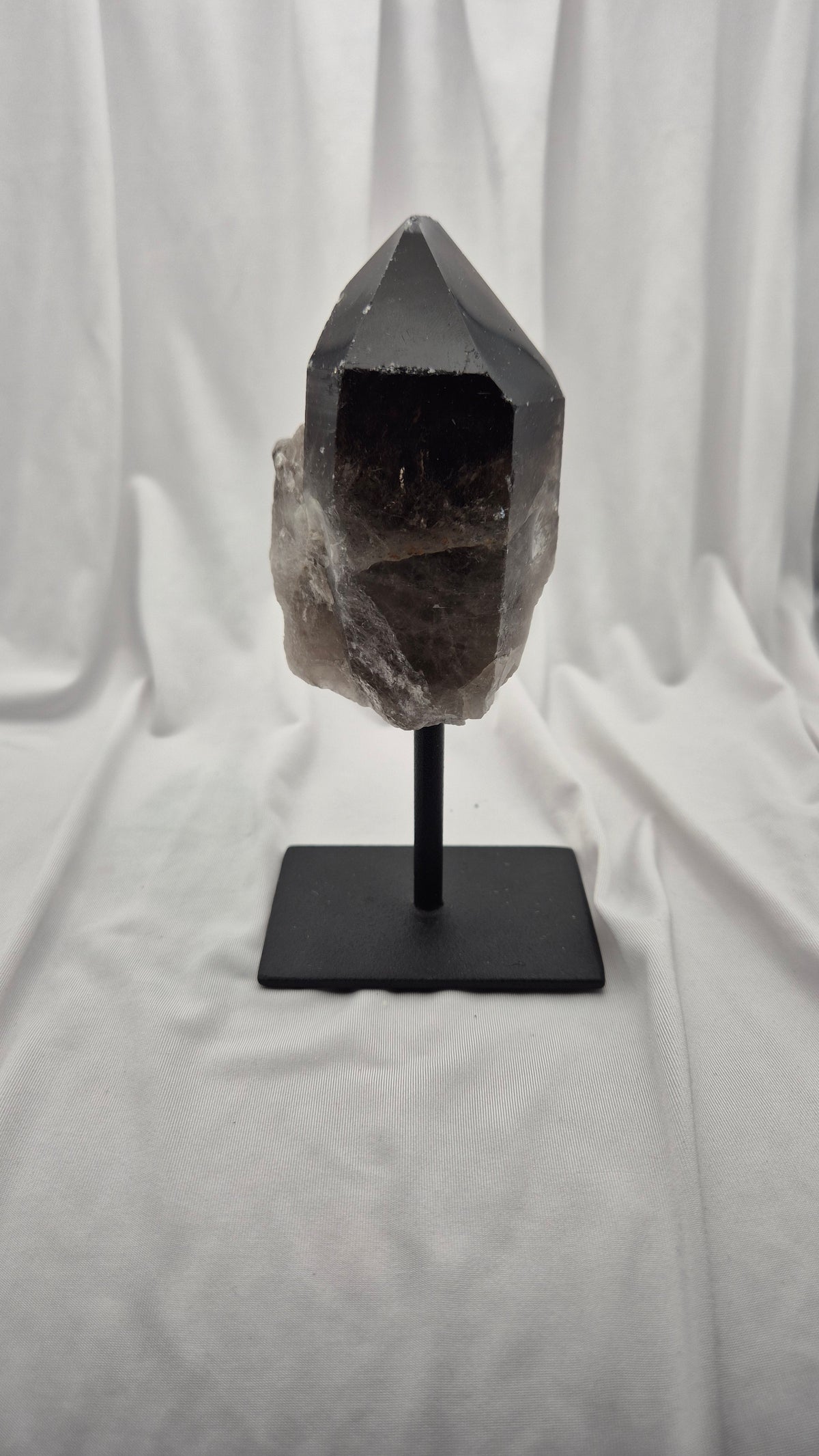 A smoky quartz crystal cluster on a black metal display stand, set against a draped white fabric background.