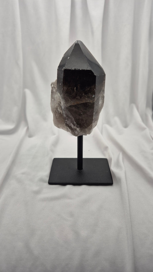 A smoky quartz crystal cluster on a black metal display stand, set against a draped white fabric background.