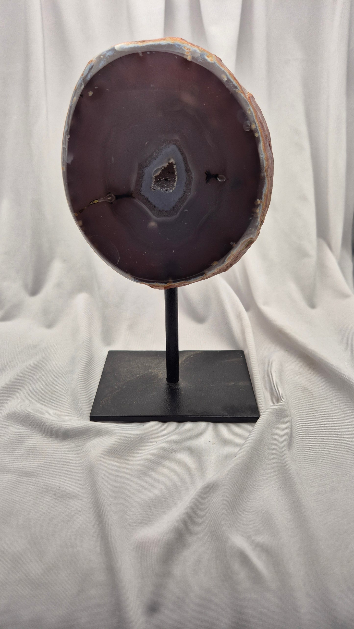 A polished agate slice display stand featuring a dark purple-brown agate slab on a black metal stand, set against a draped white fabric background.