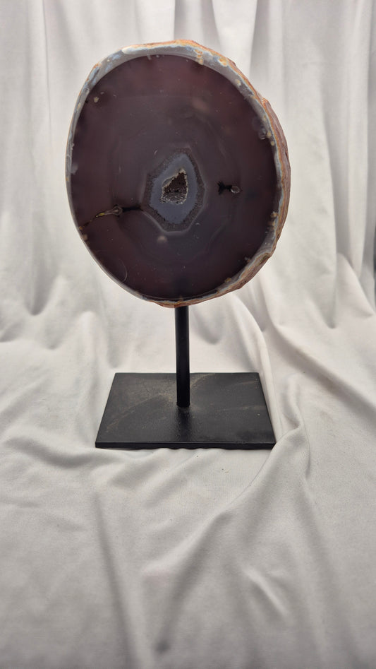 A polished agate slice display stand featuring a dark purple-brown agate slab on a black metal stand, set against a draped white fabric background.