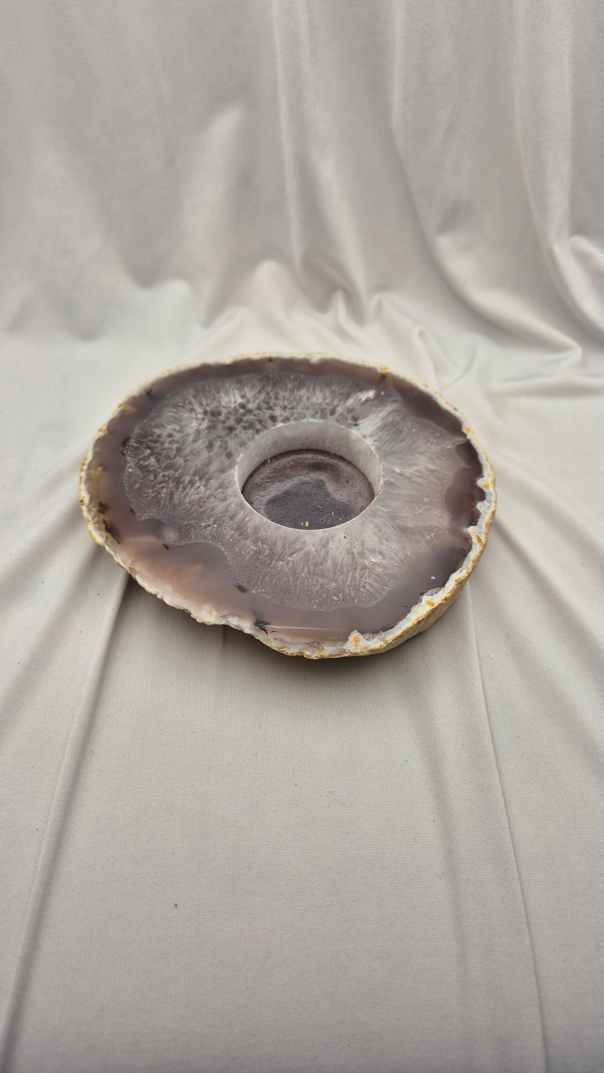 A circular agate stone bowl with a natural geode center, featuring concentric bands of gray, purple, and brown, displayed on draped white fabric.