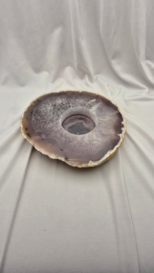 A circular agate stone bowl with a natural geode center, featuring concentric bands of gray, purple, and brown, displayed on draped white fabric.