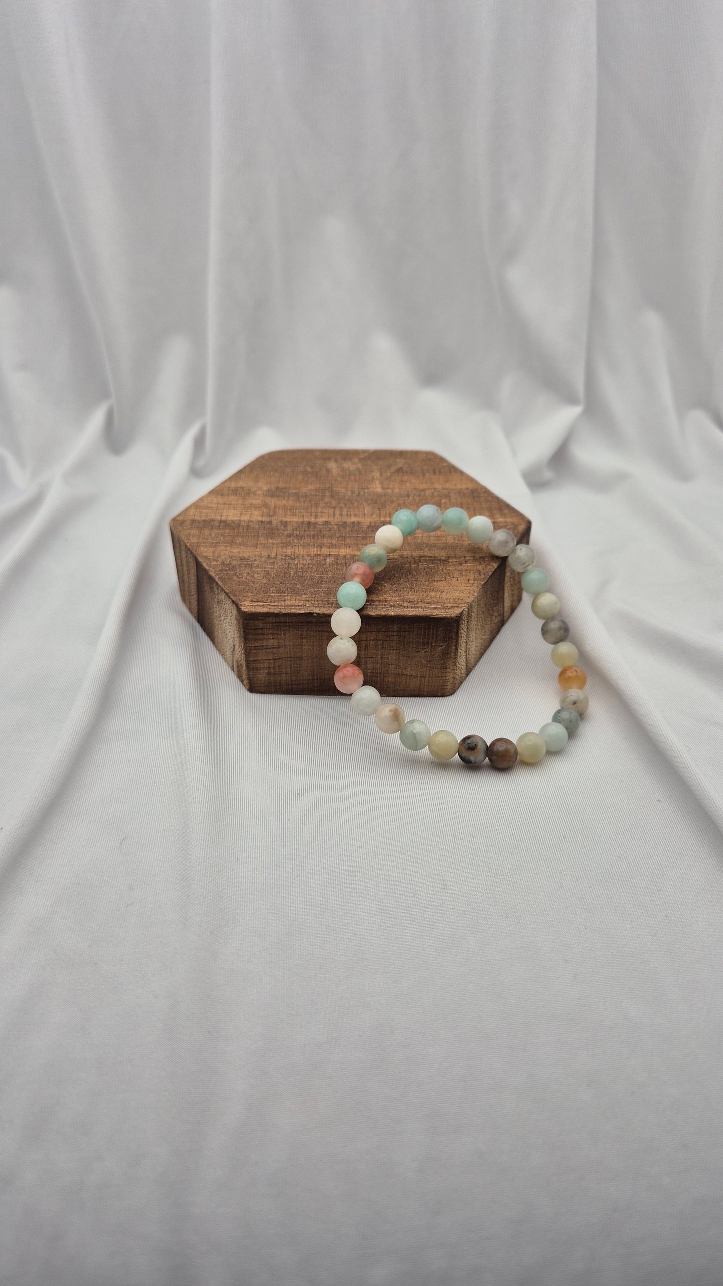 A beaded stretch bracelet made of pastel and earthy round beads, displayed on a small hexagonal wooden box against a draped white fabric backdrop.