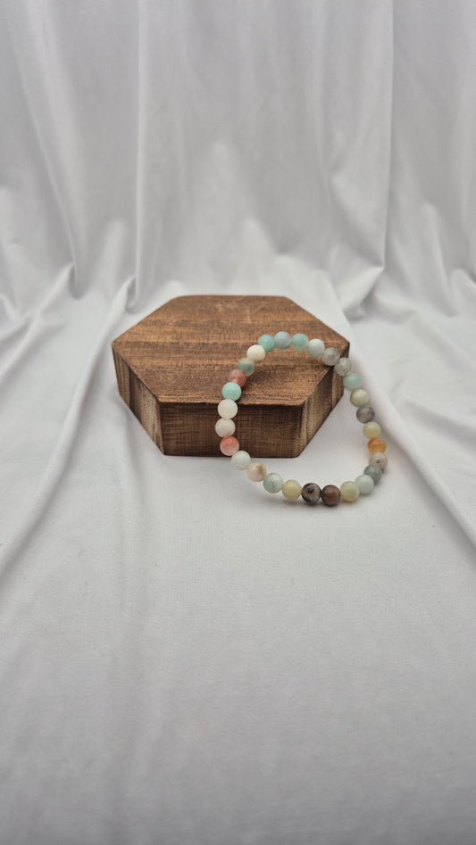A beaded stretch bracelet made of pastel and earthy round beads, displayed on a small hexagonal wooden box against a draped white fabric backdrop.