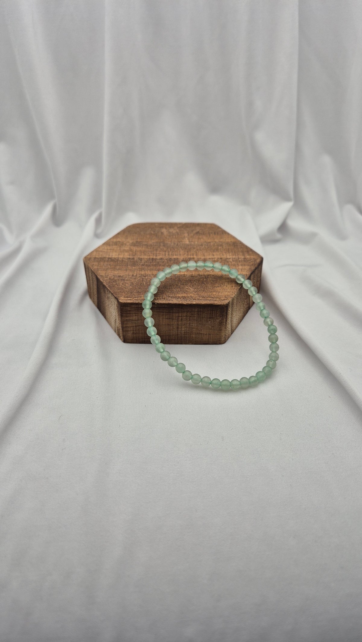 A light green beaded bracelet resting on a hexagonal wooden block against a draped white fabric backdrop.
