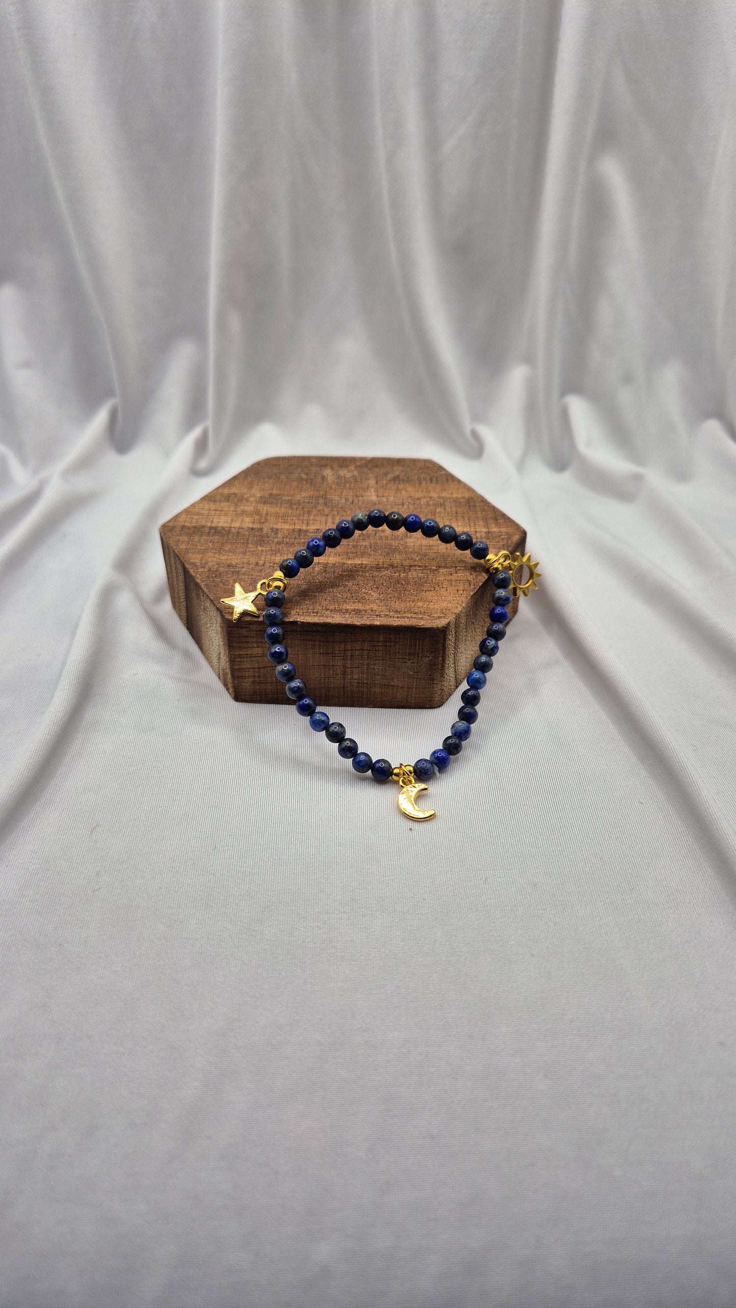 A beaded bracelet with dark blue beads and gold-toned celestial charms, displayed on a small wooden block against a draped light gray fabric backdrop.
