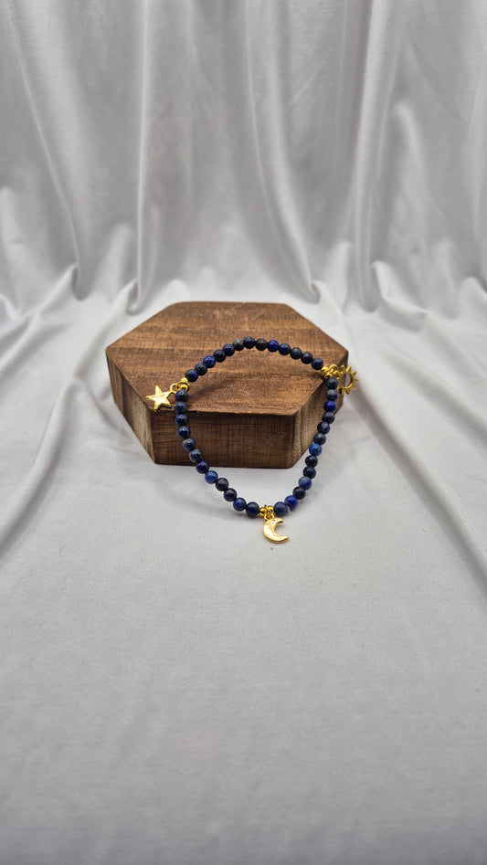 A beaded bracelet with dark blue beads and gold-toned celestial charms, displayed on a small wooden block against a draped light gray fabric backdrop.