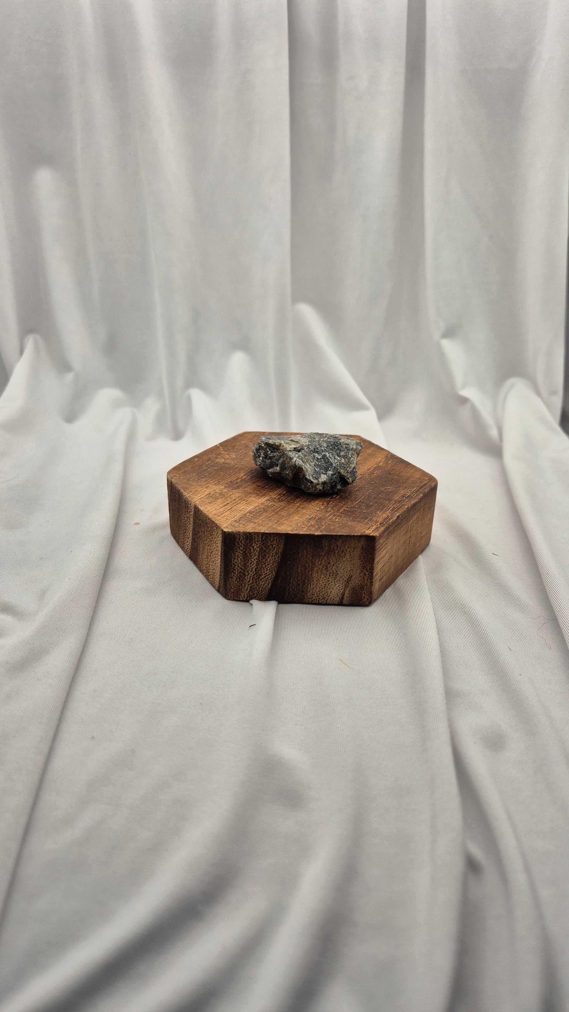 A small hexagonal wooden box with a rough dark gray stone or crystal resting on top, set against a draped white fabric backdrop.