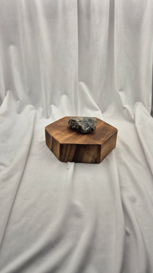 A small hexagonal wooden box with a rough dark gray stone or crystal resting on top, set against a draped white fabric backdrop.