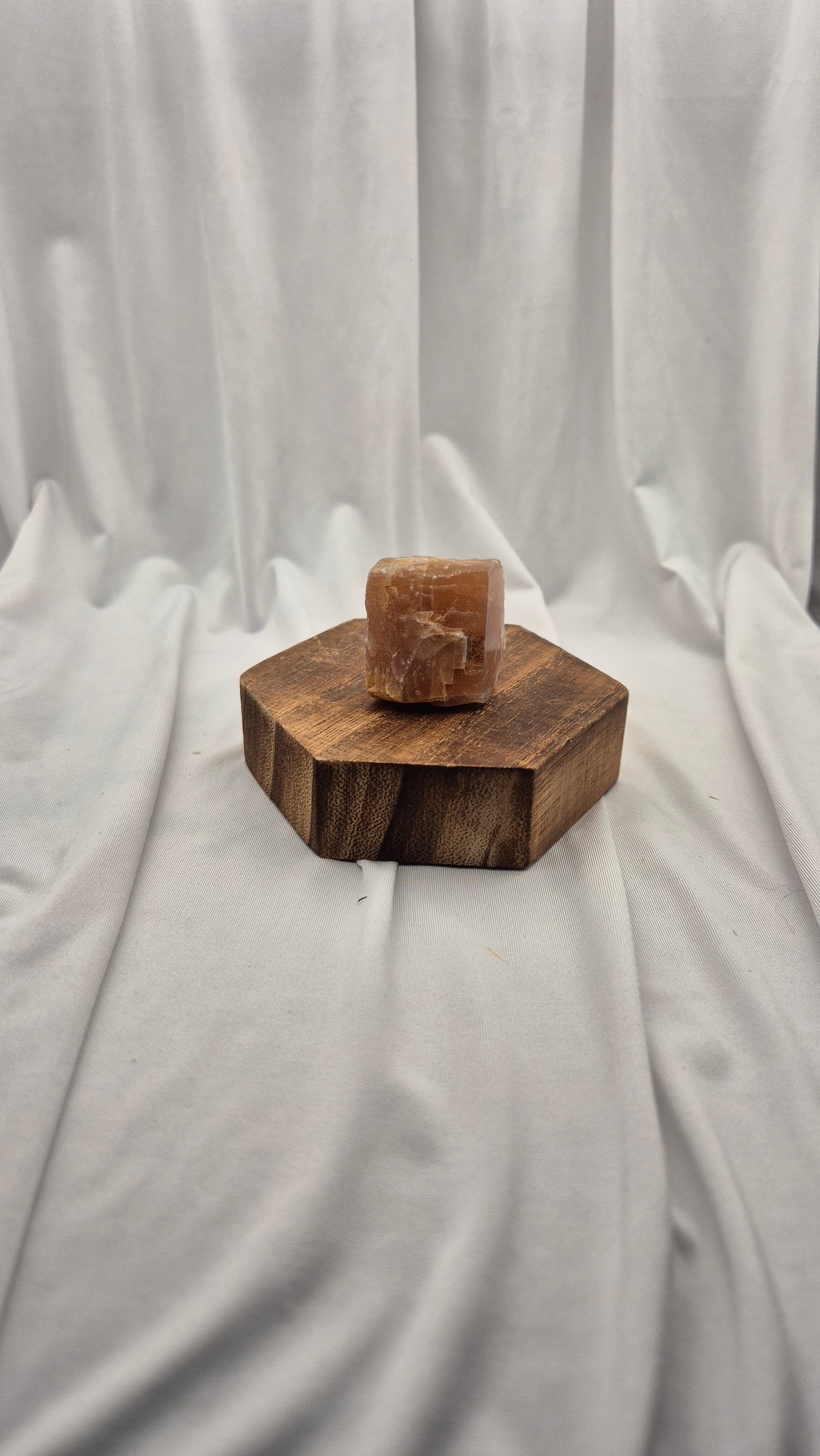 A polished rectangular stone sample in warm tan-brown sits centered on a hexagonal wooden block, set against draped white fabric.
