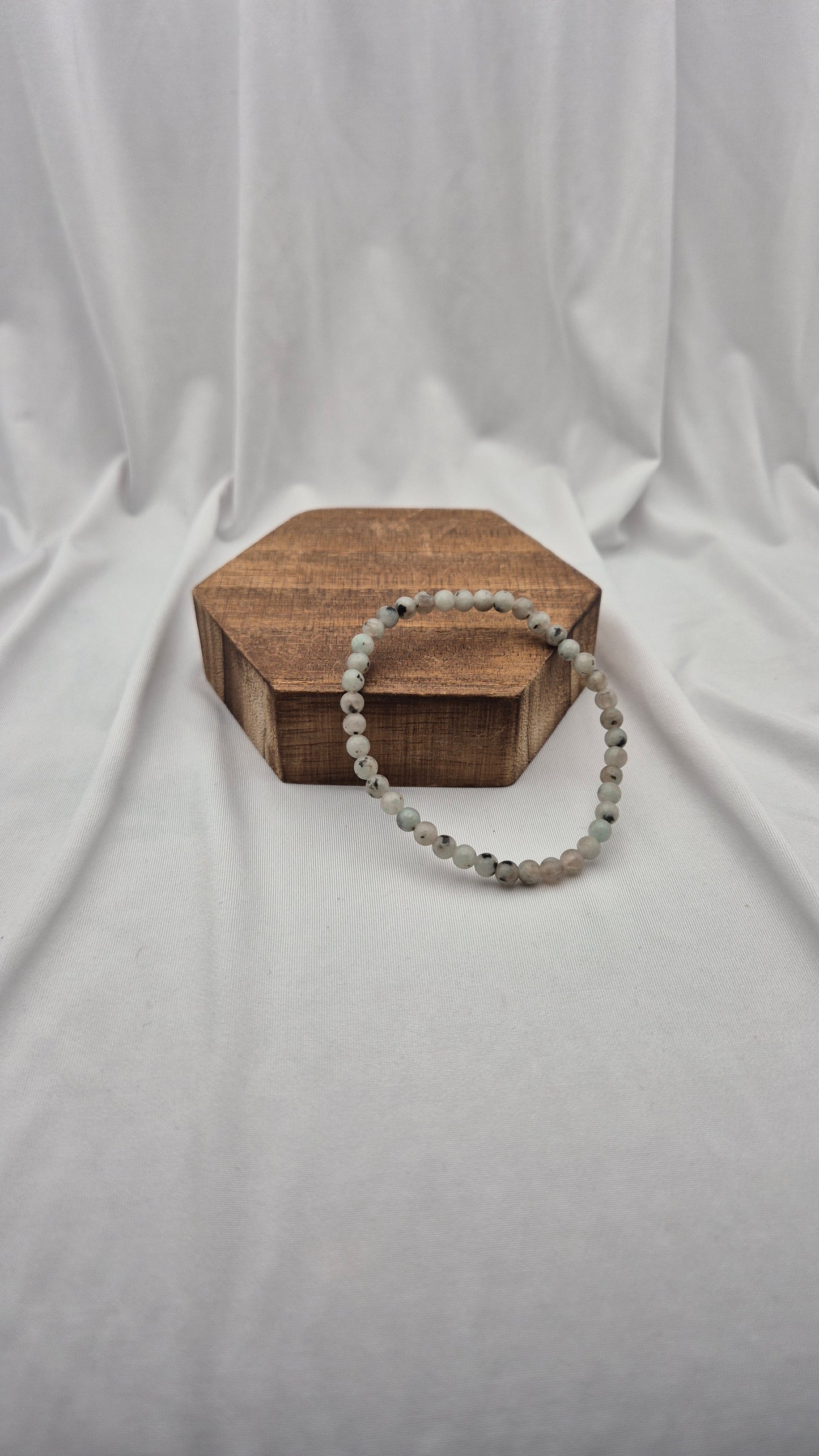 A beaded stretch bracelet made of light gray and white marbled beads, draped over a small hexagonal wooden box on a draped white fabric background.