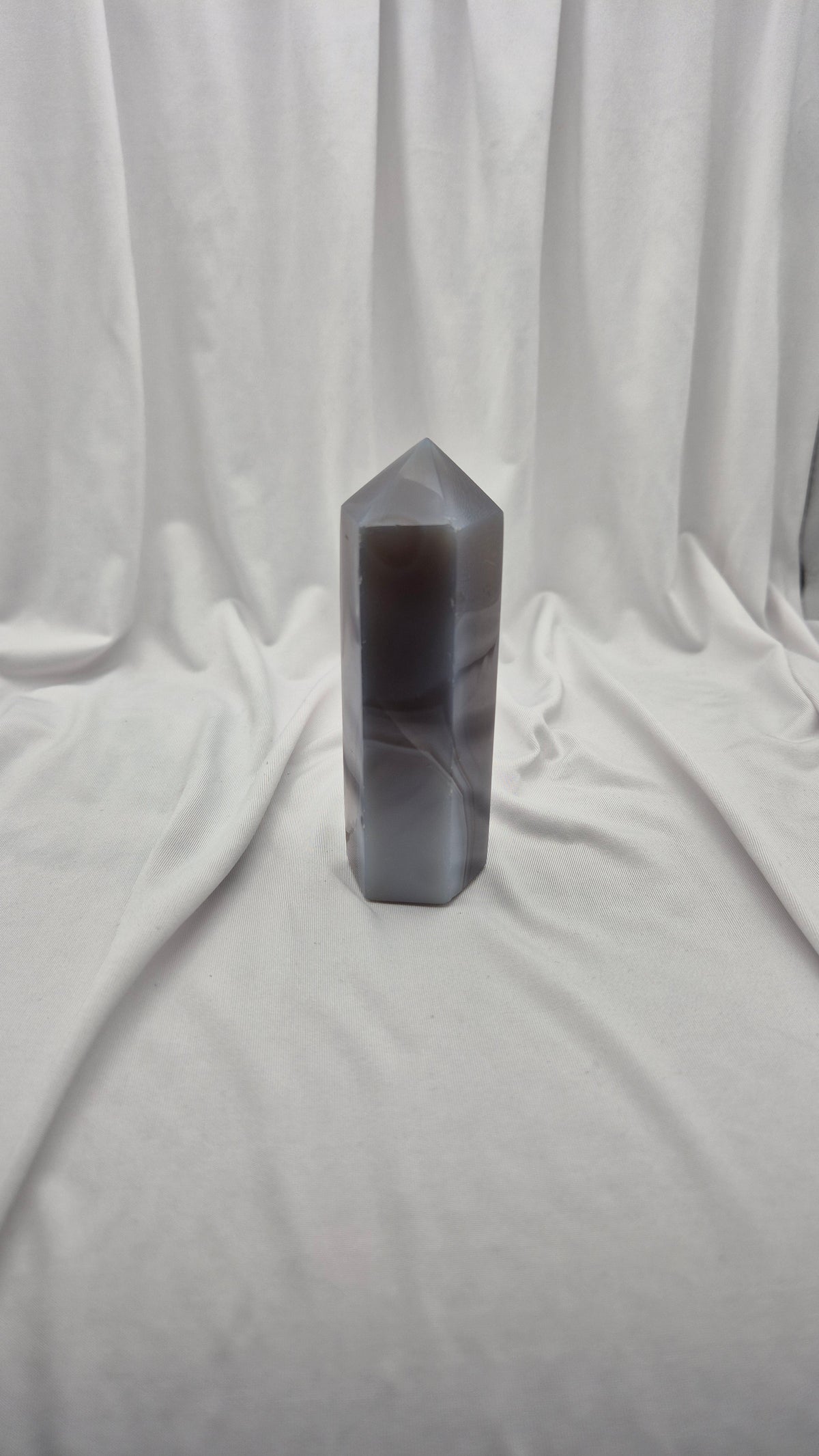 A tall, hexagonal polished crystal tower with a pointed top, made of semi-translucent gray agate, standing on a draped white fabric backdrop.