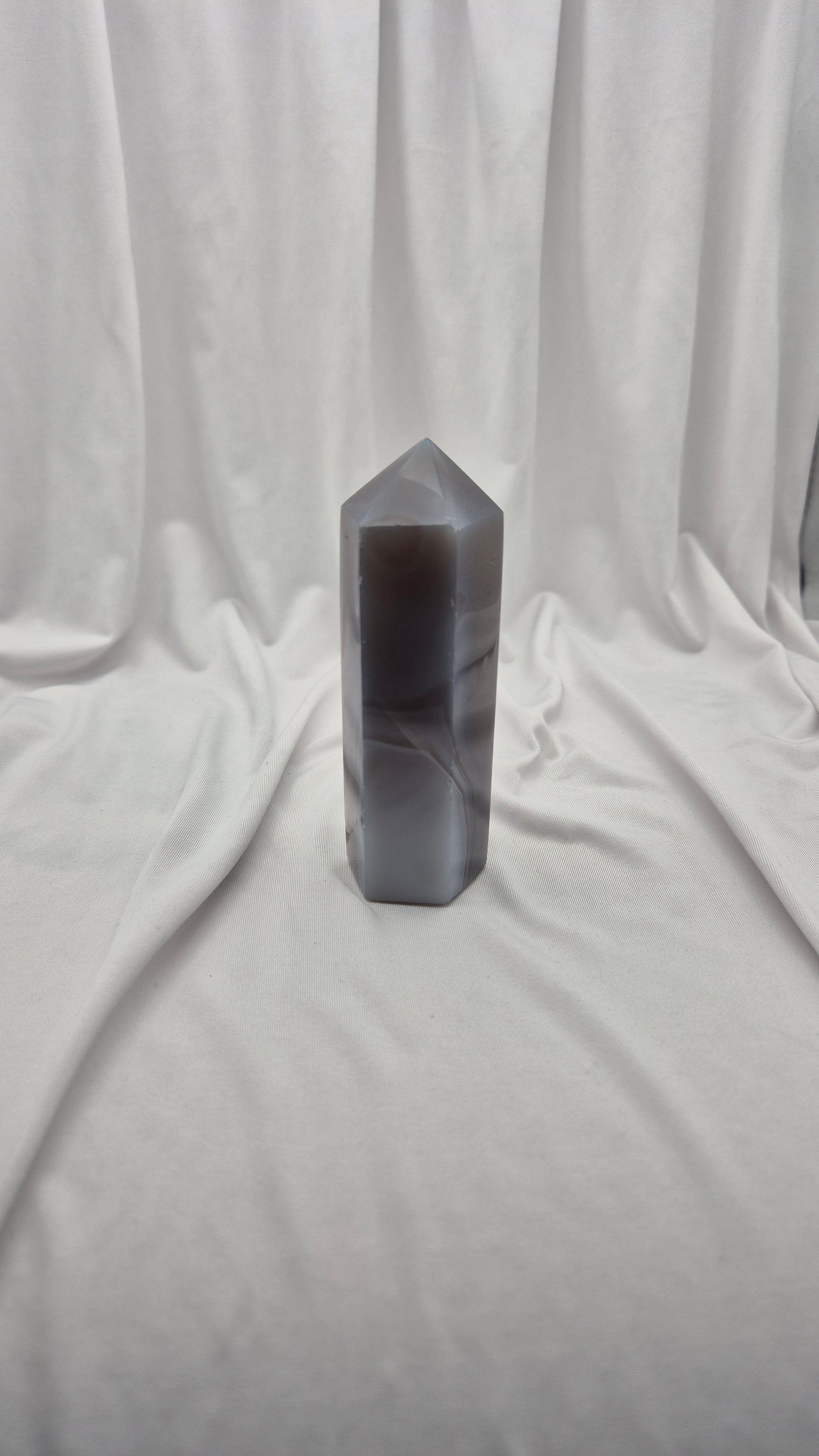 A tall, hexagonal polished crystal tower with a pointed top, made of semi-translucent gray agate, standing on a draped white fabric backdrop.