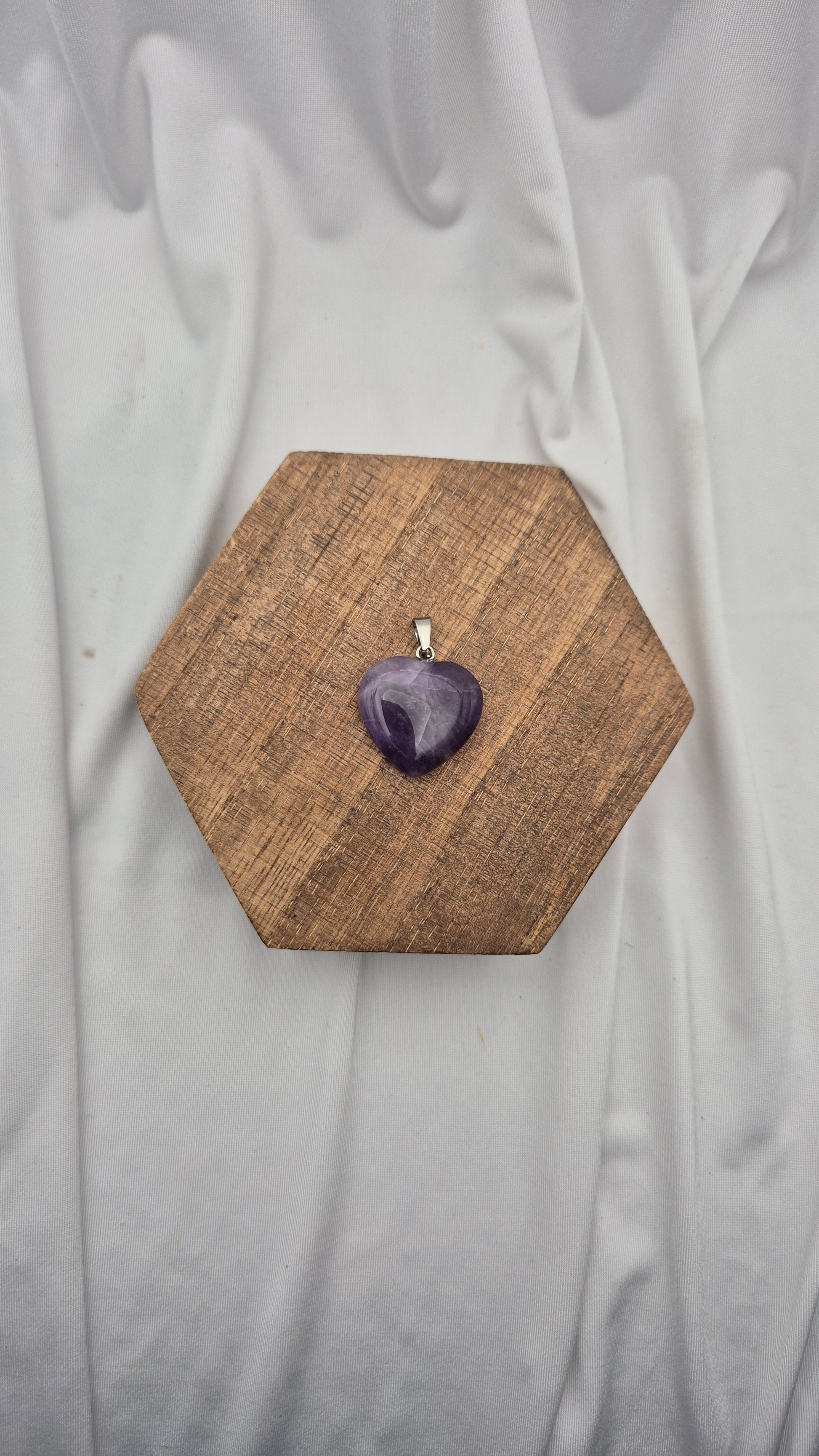 A heart-shaped purple amethyst gemstone pendant on a silver bail, displayed on a hexagonal wooden block over a draped white fabric background.