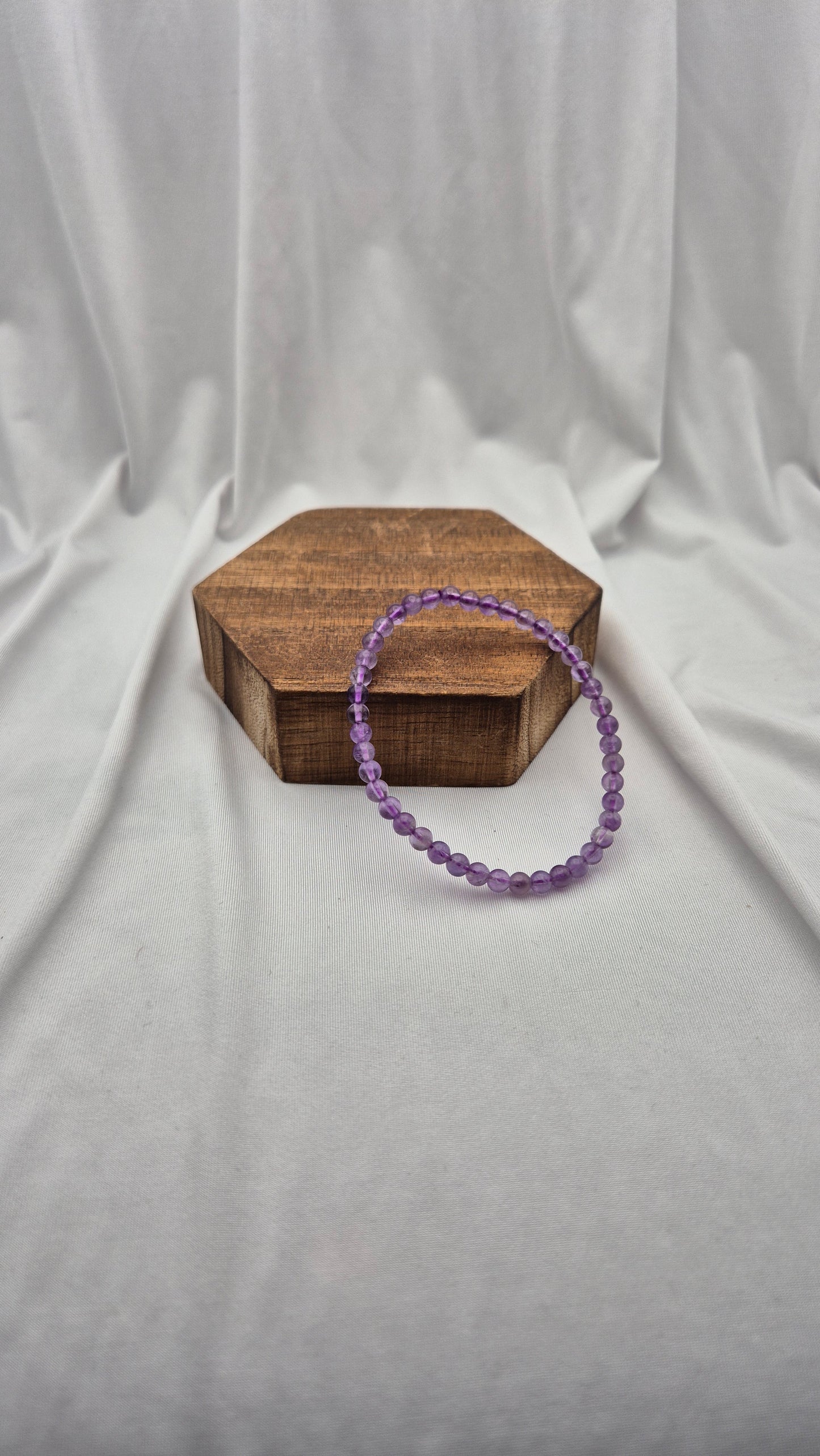 A purple beaded bracelet draped over a small hexagonal wooden box, set against a draped white fabric backdrop.