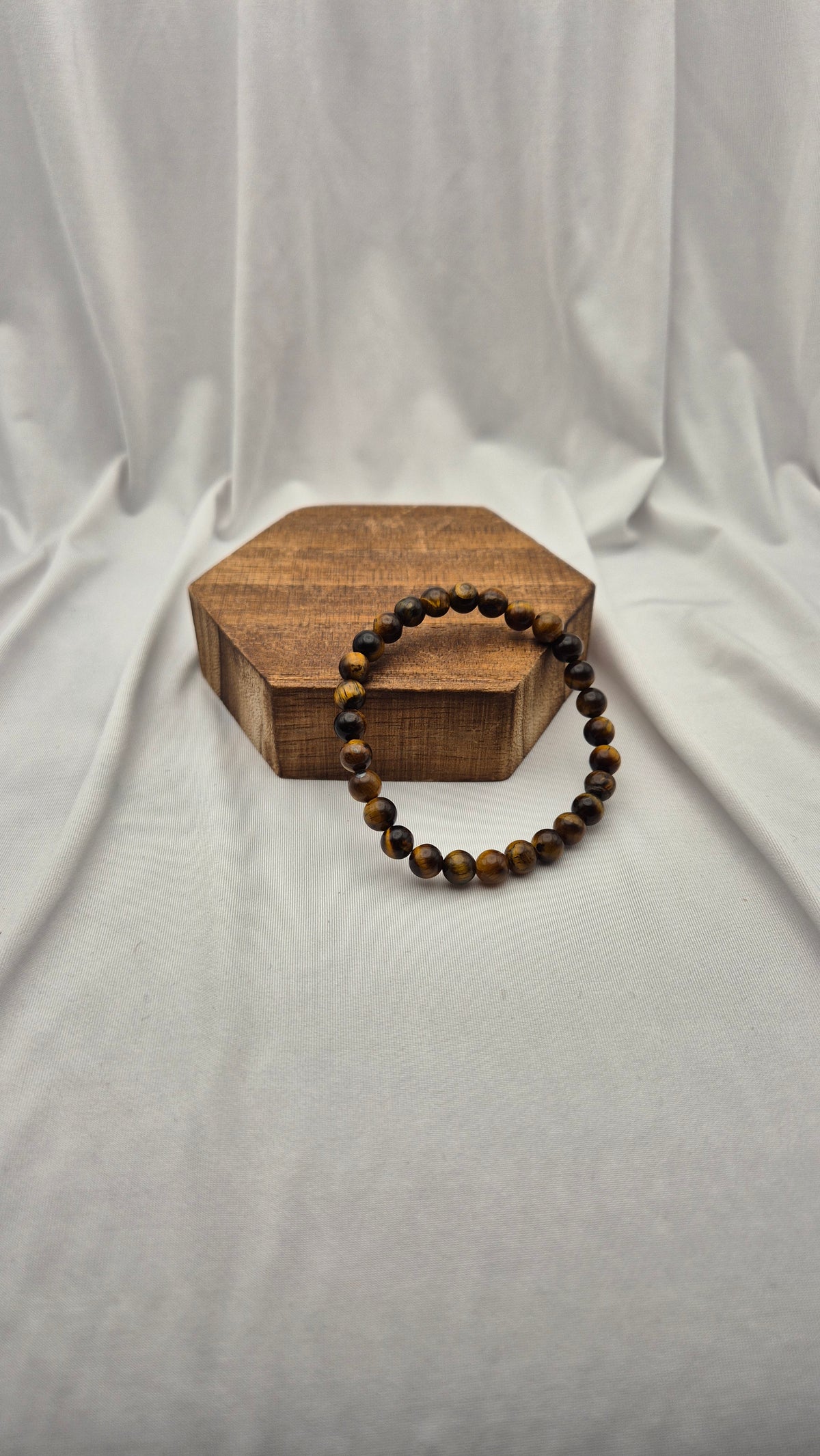 A beaded bracelet made of brown and black tiger's eye beads, displayed on a small hexagonal wooden box against a draped white fabric background.