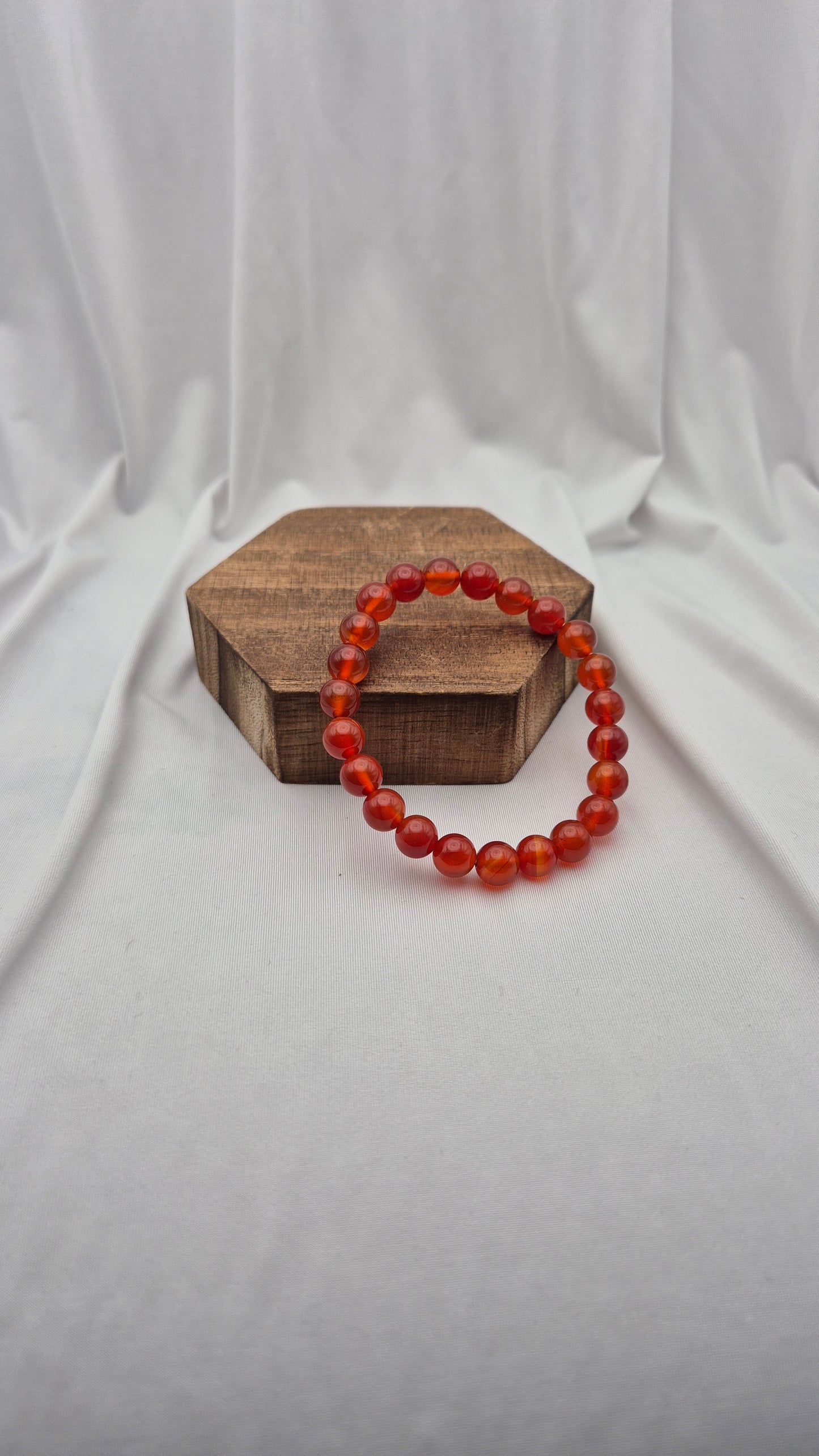 A beaded stretch bracelet made of translucent orange-red round beads, displayed on a small hexagonal wooden block against a draped white fabric backdrop.