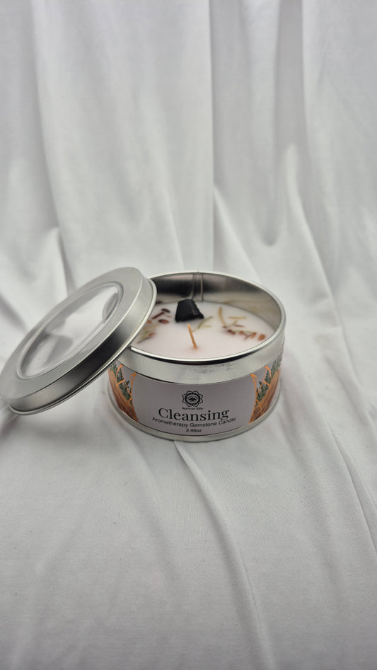 A silver metal tin candle labeled 'Cleansing' with a white wax surface and dried botanical pieces, shown with its lid off on a draped white fabric background.