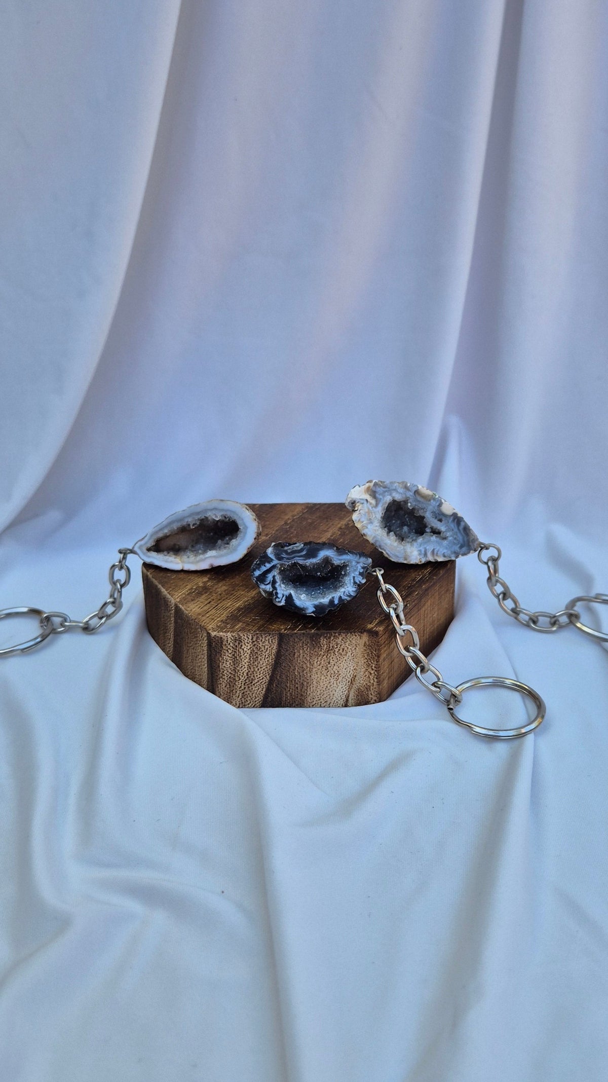 Three silver chain keychains with natural agate geode pendants rest on a small wooden block against a draped white fabric backdrop.