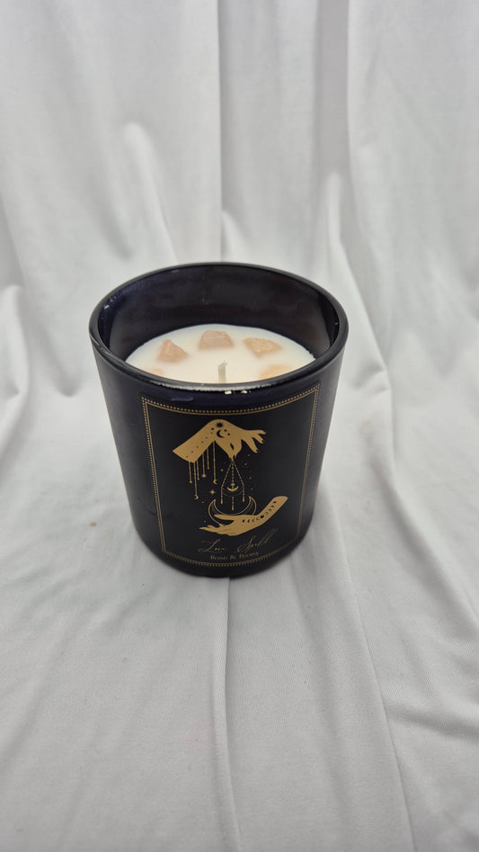 A black glass jar candle with a gold illustration of a hand holding a dripping bell and a wick at the top, filled with white wax and set against a draped white fabric background.