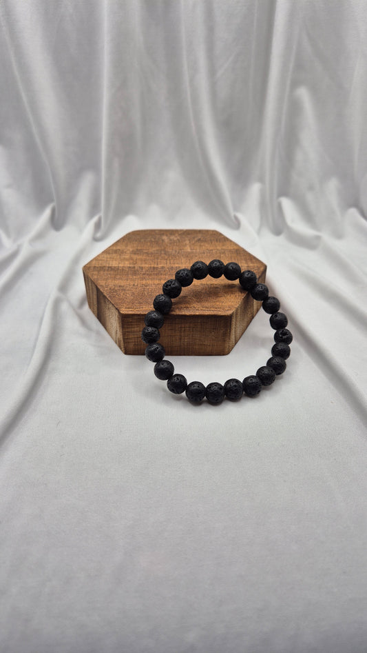 A black beaded bracelet resting on a small hexagonal wooden block, set against a draped white fabric backdrop.