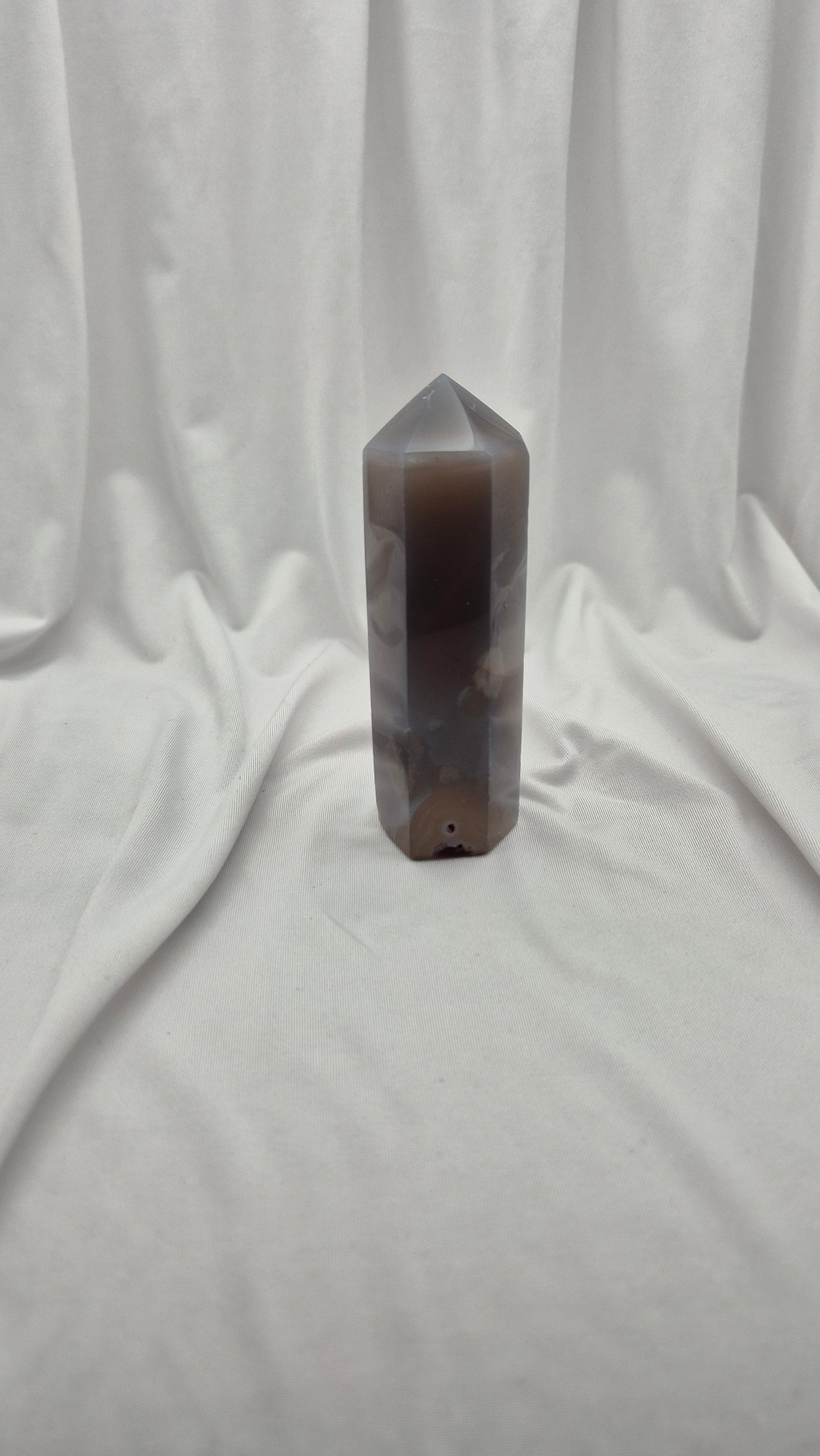 A polished brown and gray agate crystal tower with a pointed tip, standing upright on a draped white fabric background.