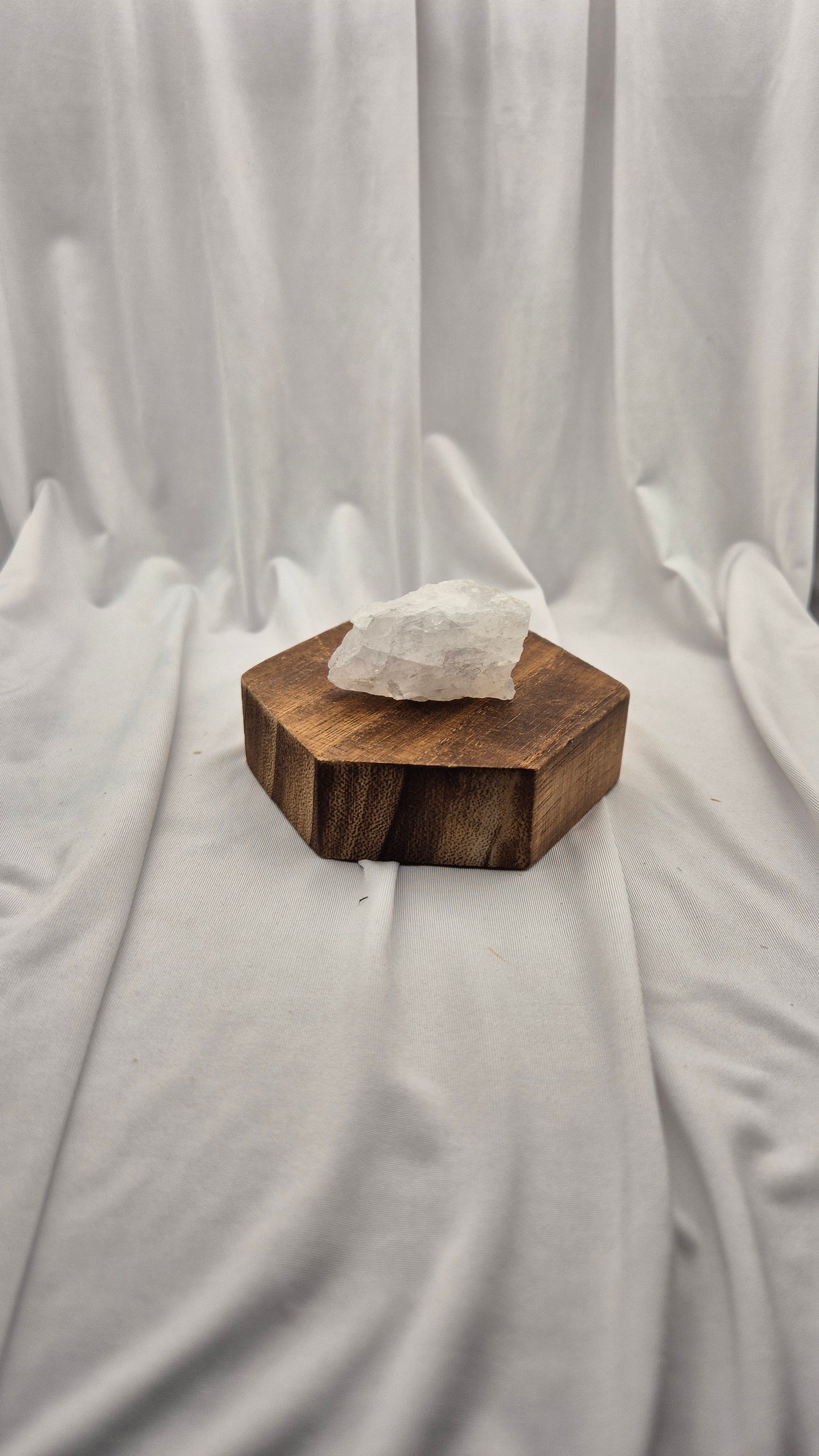 A rough white crystal stone resting on a hexagonal dark brown wooden block, set against a draped white fabric backdrop.