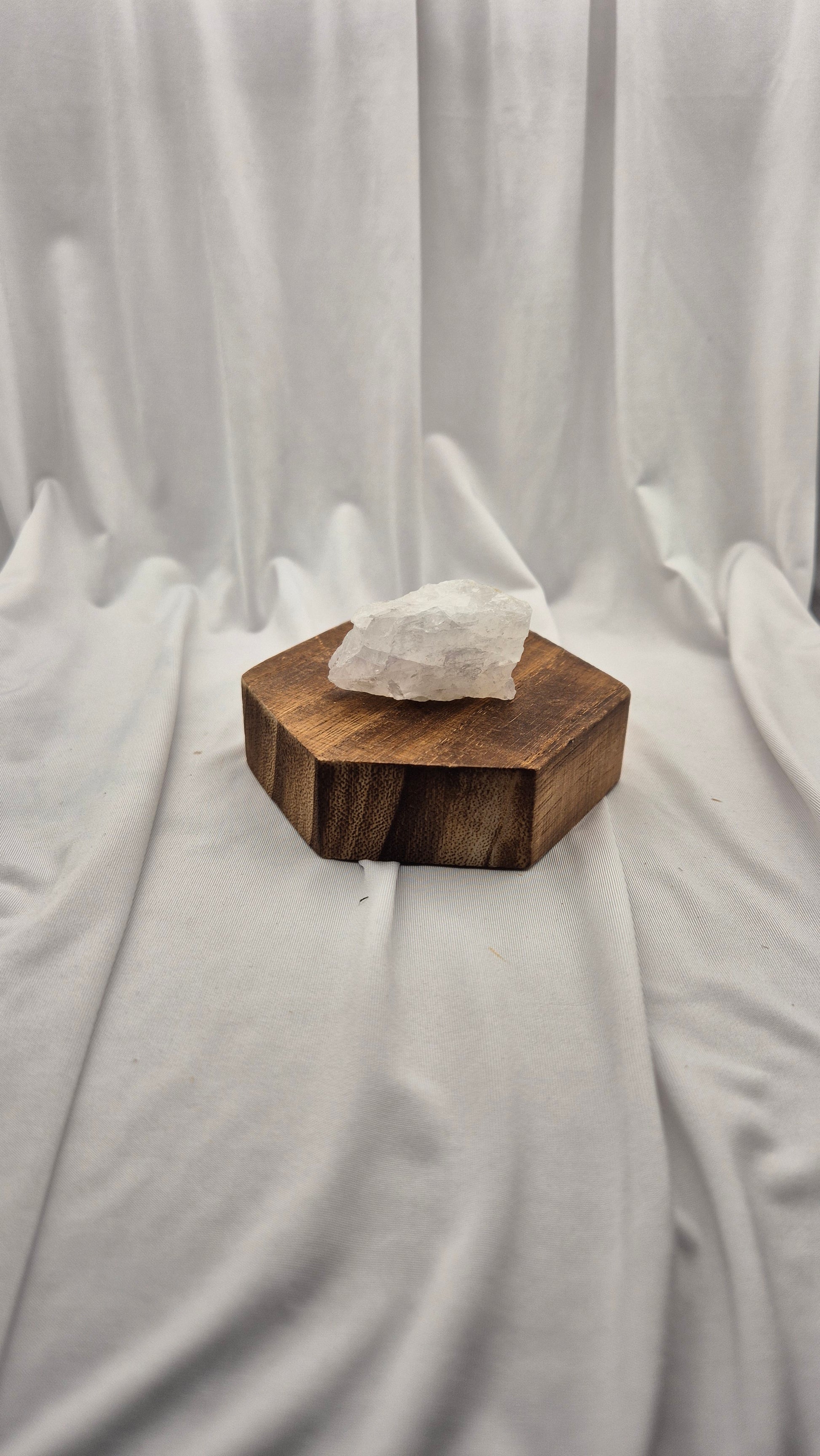 A rough white crystal stone resting on a hexagonal dark brown wooden block, set against a draped white fabric backdrop.