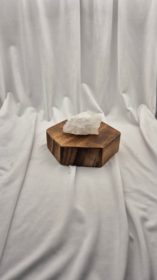 A rough white crystal stone resting on a hexagonal dark brown wooden block, set against a draped white fabric backdrop.