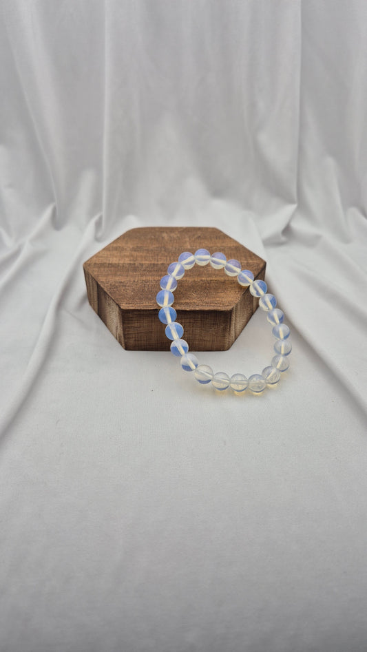 A beaded stretch bracelet made of translucent white beads with a subtle blue iridescent sheen, resting on a small hexagonal wooden box against a draped white fabric backdrop.