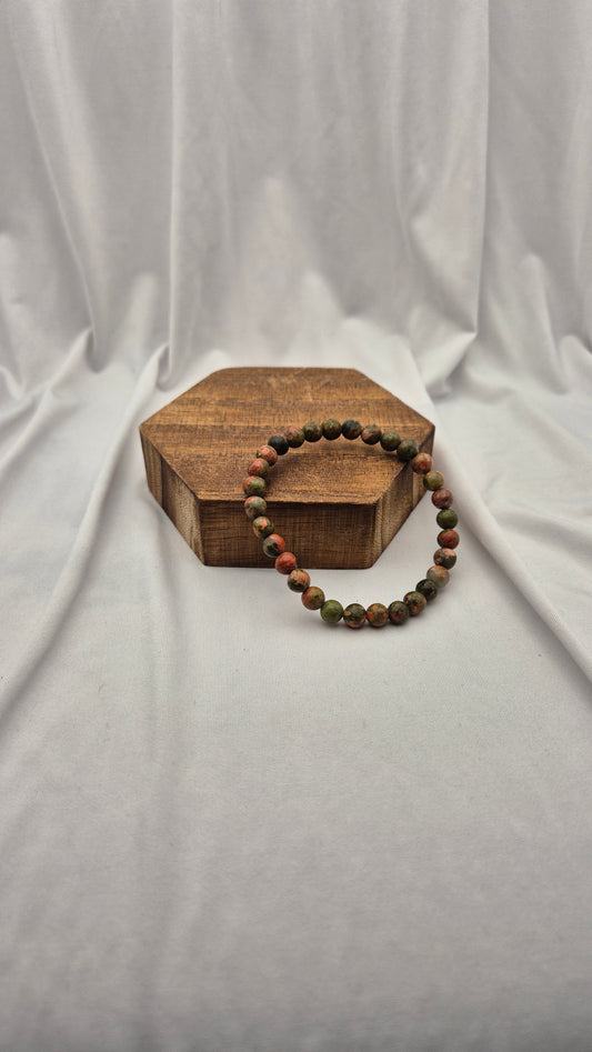 A beaded stretch bracelet with round brown and green beads, displayed on a small hexagonal wooden block against a draped white fabric backdrop.