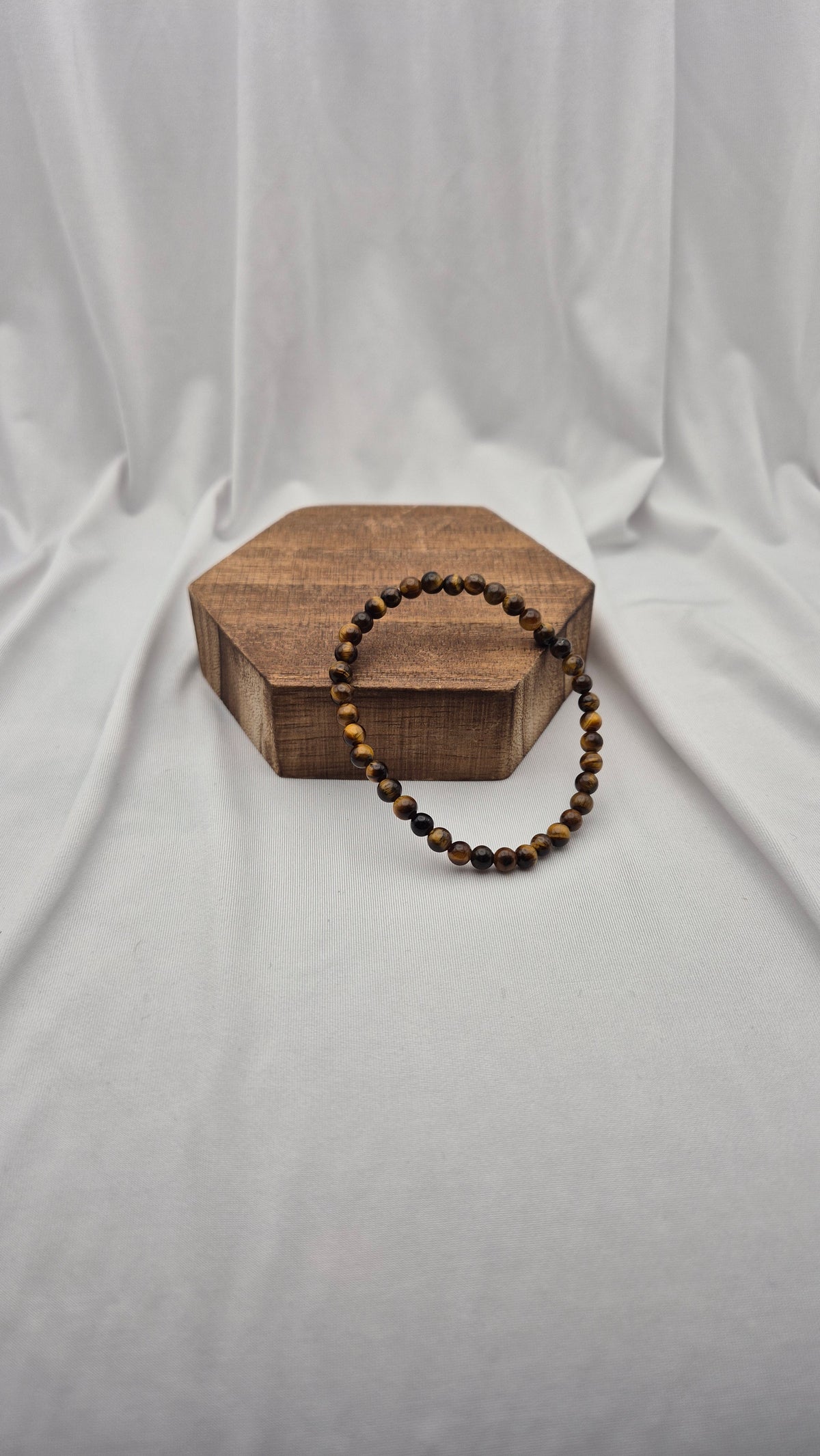 A beaded bracelet with brown and black tiger's eye beads draped over a small hexagonal wooden box, set against a draped white fabric background.