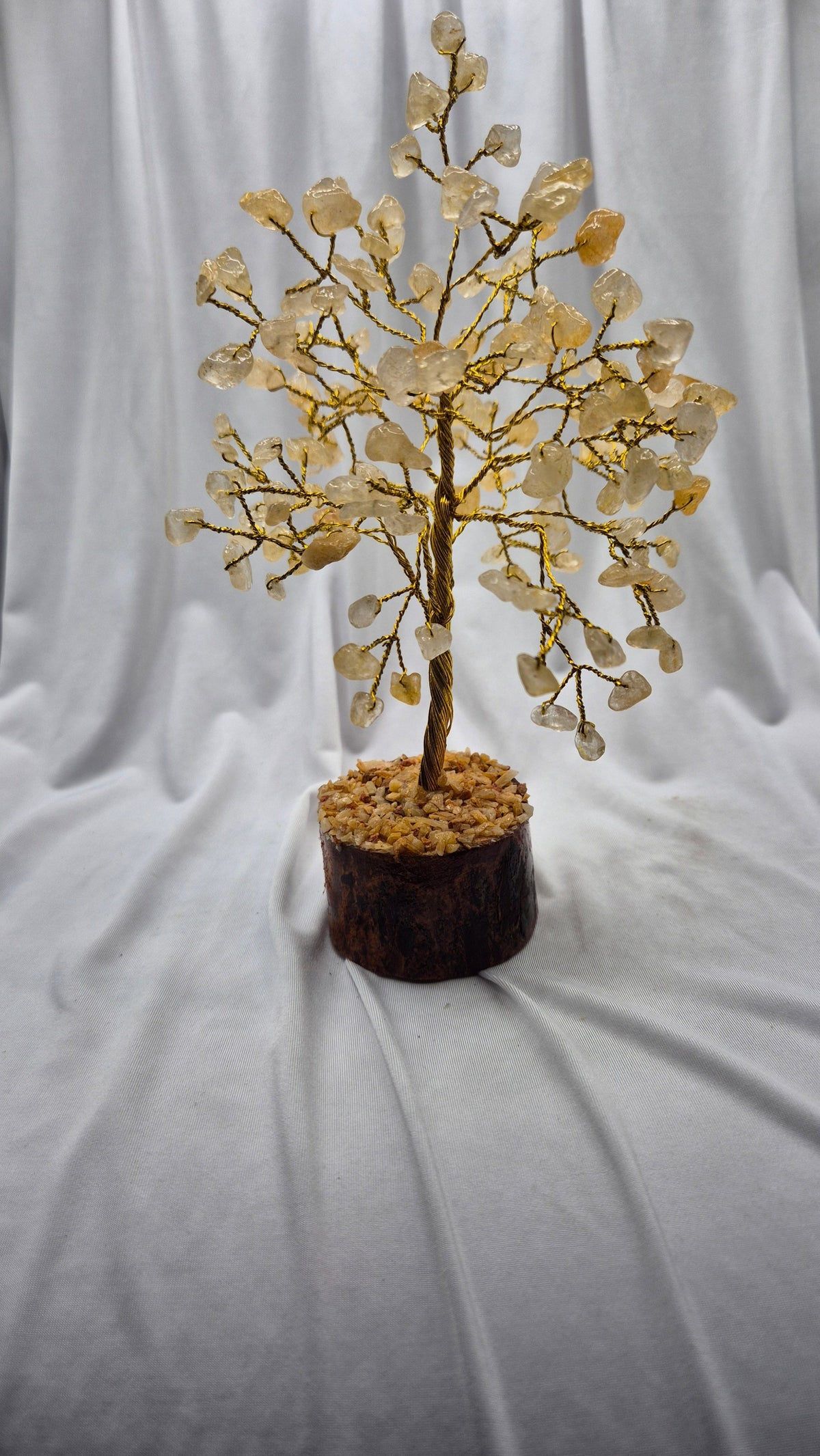 A crystal tree decorative piece with gold-toned branches and pale yellow quartz-like leaves, displayed in a dark brown wooden pot filled with small pebbles, set against a draped white fabric backdrop.