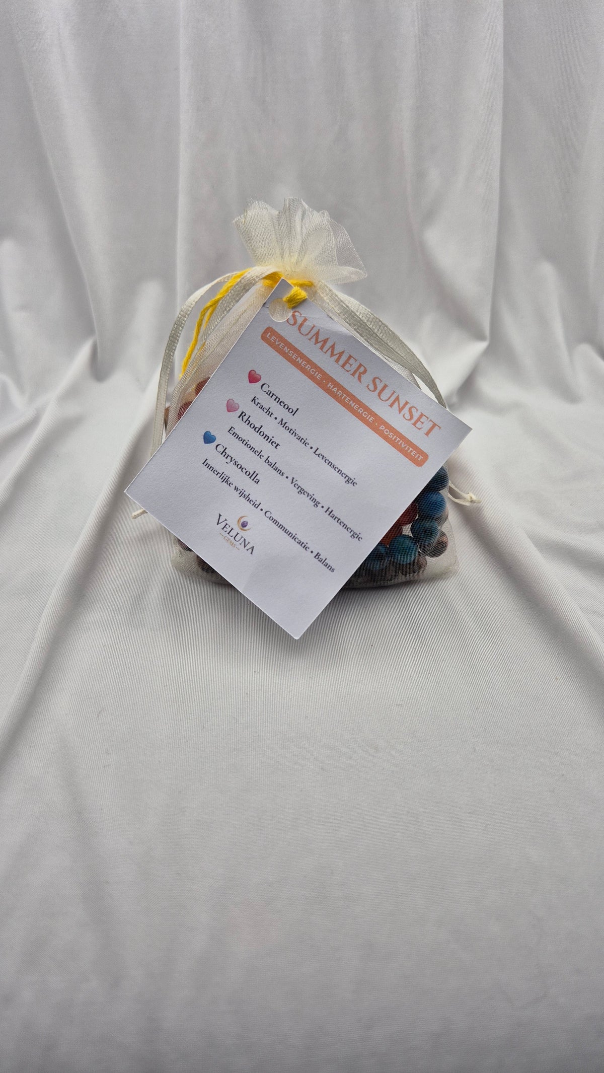A small gift bag tied with a sheer white organza bow, containing a packaged item and a printed tag that reads 'SUMMER SUNSET' and 'Vilena', placed on a white fabric backdrop.