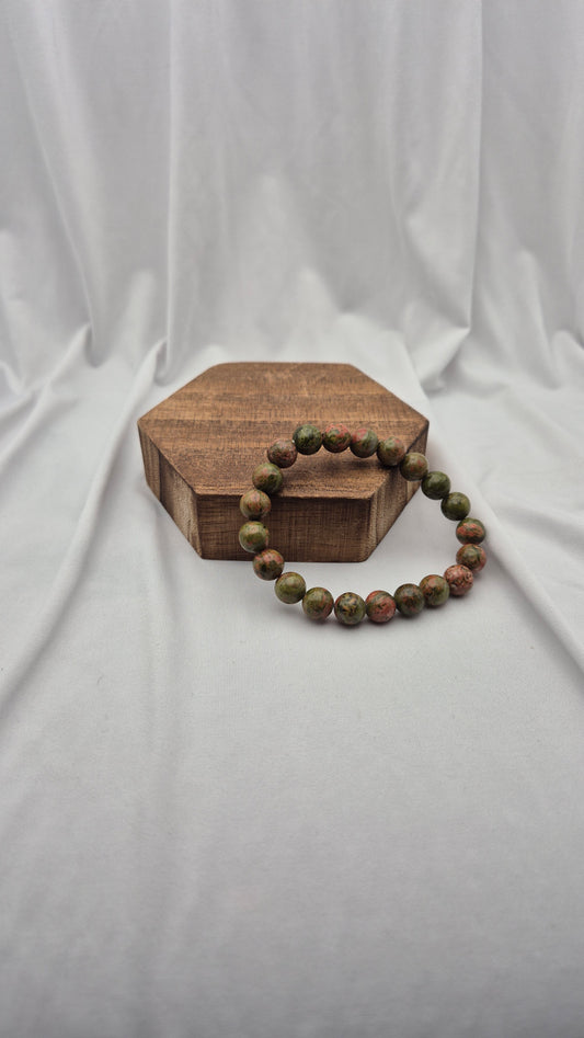 A beaded bracelet made of round brown and green stones, displayed on a small hexagonal wooden block against a draped white fabric backdrop.