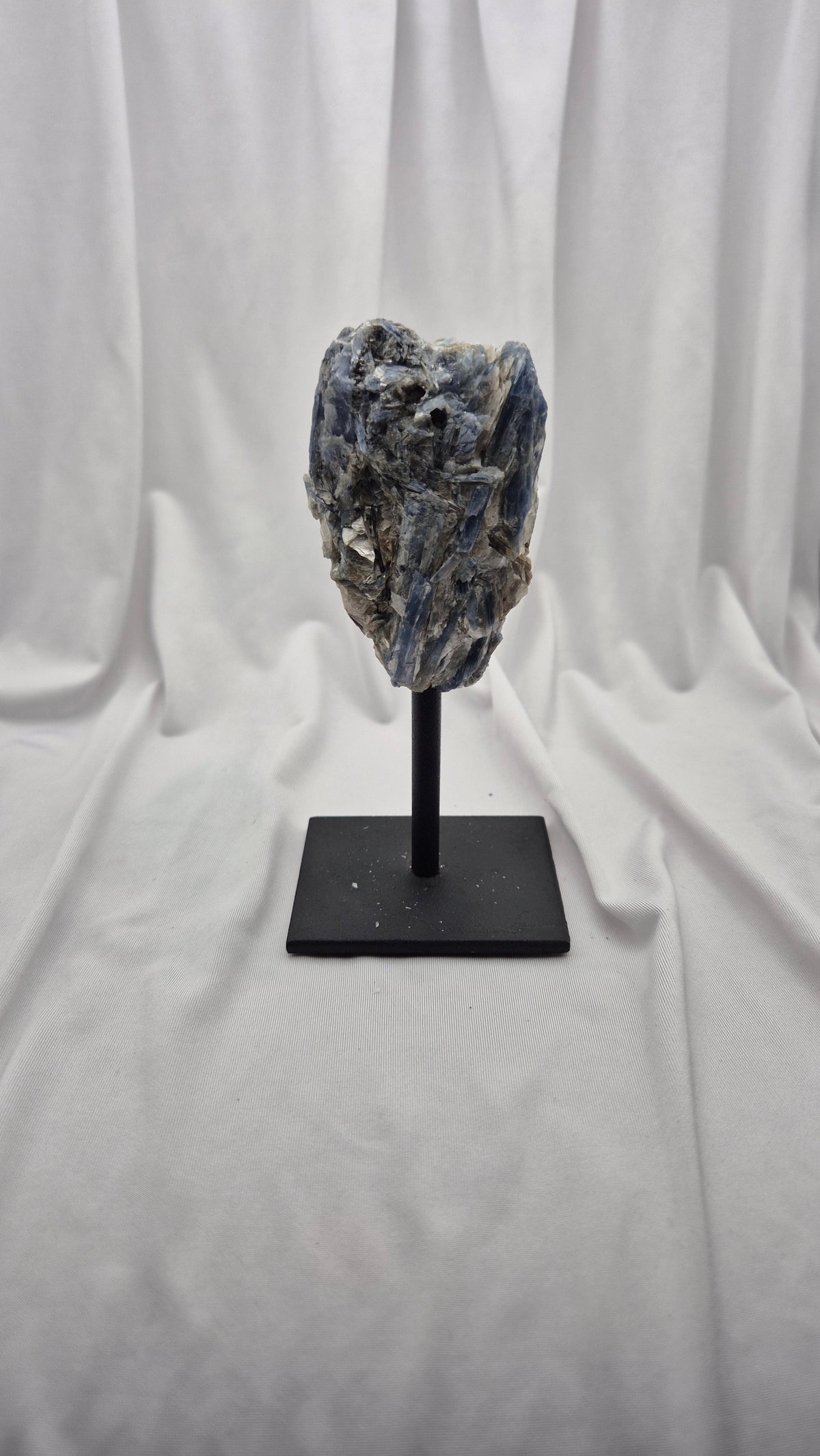 A natural blue-gray mineral specimen or crystal cluster mounted on a black display stand against a draped white fabric backdrop.