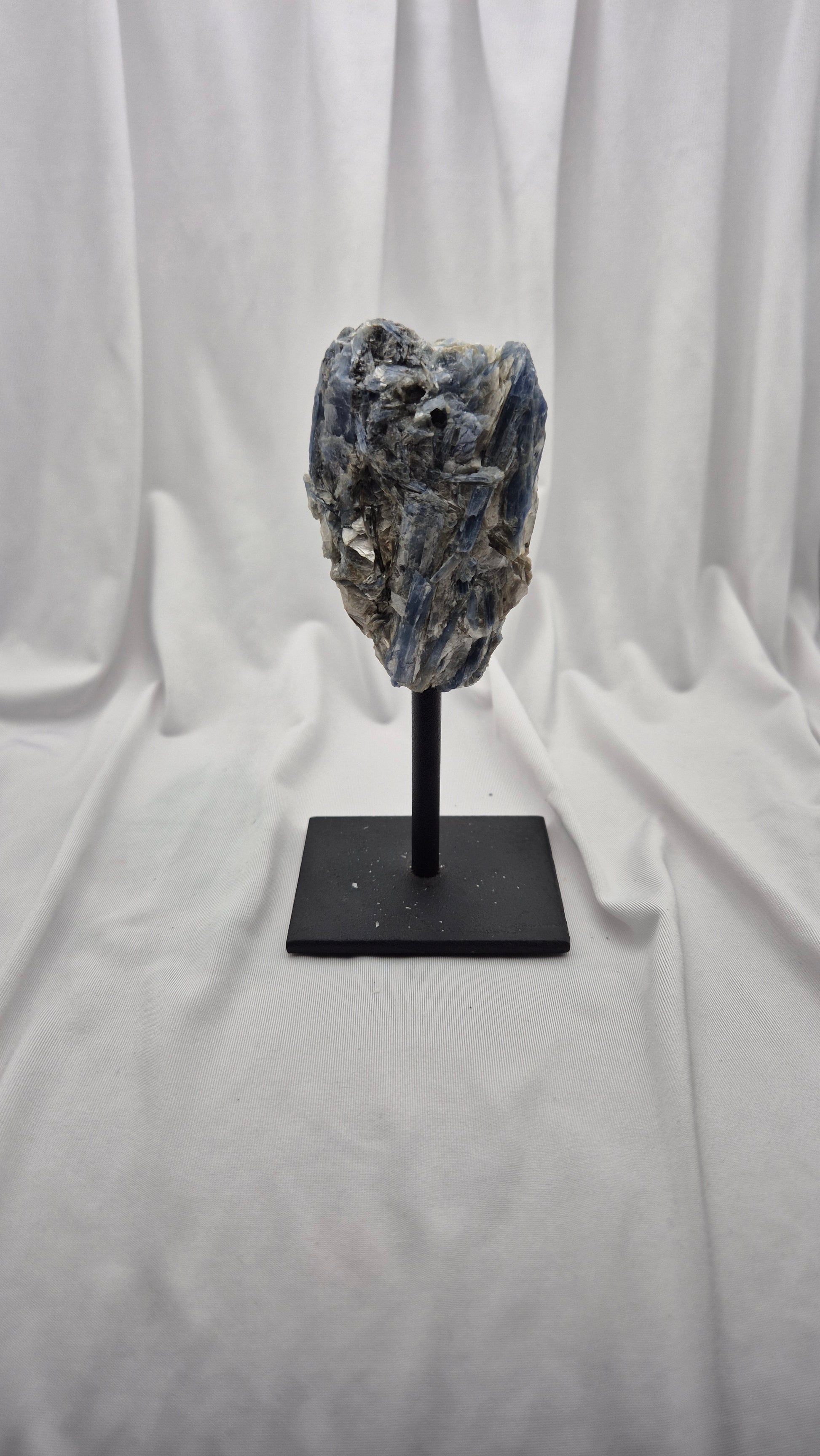A natural blue-gray mineral specimen or crystal cluster mounted on a black display stand against a draped white fabric backdrop.