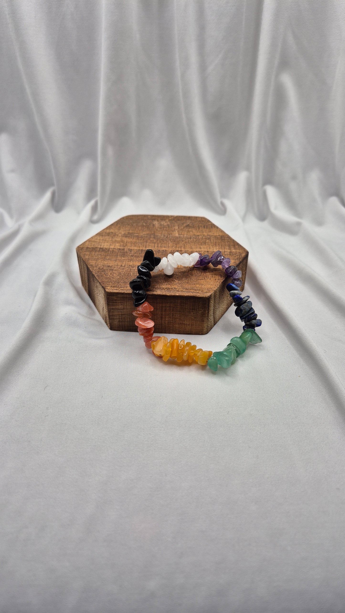 A multicolor chakra gemstone bead bracelet displayed on a small hexagonal wooden block against a draped white fabric background.