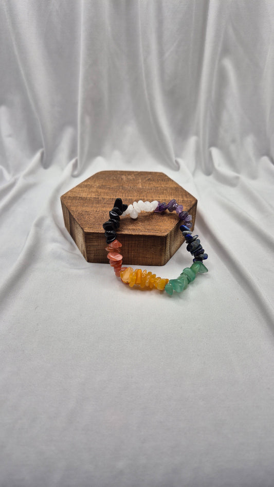 A multicolor chakra gemstone bead bracelet displayed on a small hexagonal wooden block against a draped white fabric background.