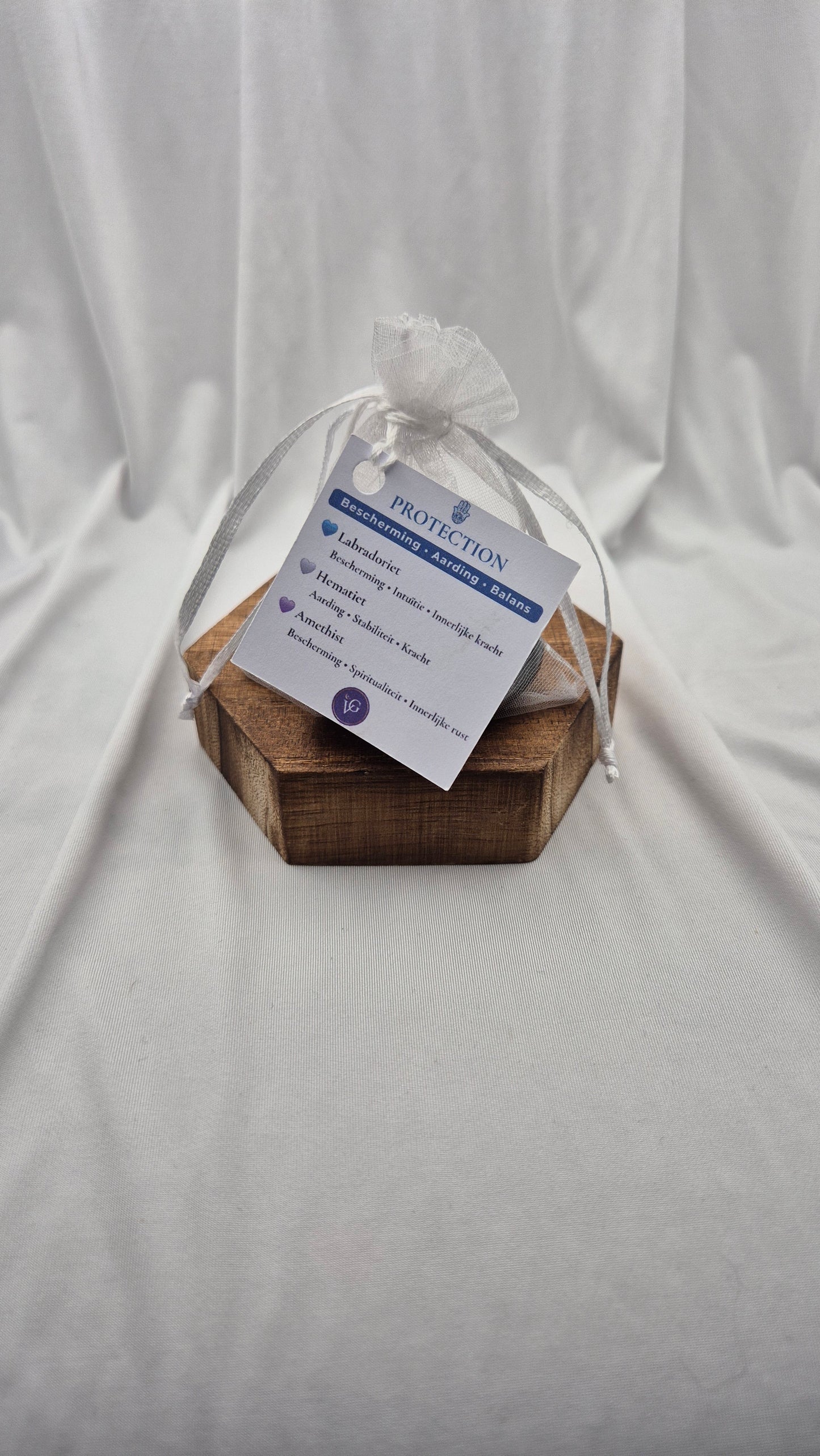 A small hexagonal wooden box wrapped in white cellophane and tied with a white ribbon, holding a printed white tag that reads 'PROTECTION' and 'BREATHE EASY'. The box is placed on a draped white fabric background.