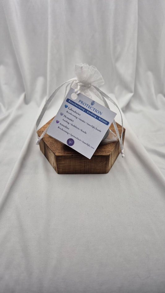 A small hexagonal wooden box wrapped in white cellophane and tied with a white ribbon, holding a printed white tag that reads 'PROTECTION' and 'BREATHE EASY'. The box is placed on a draped white fabric background.