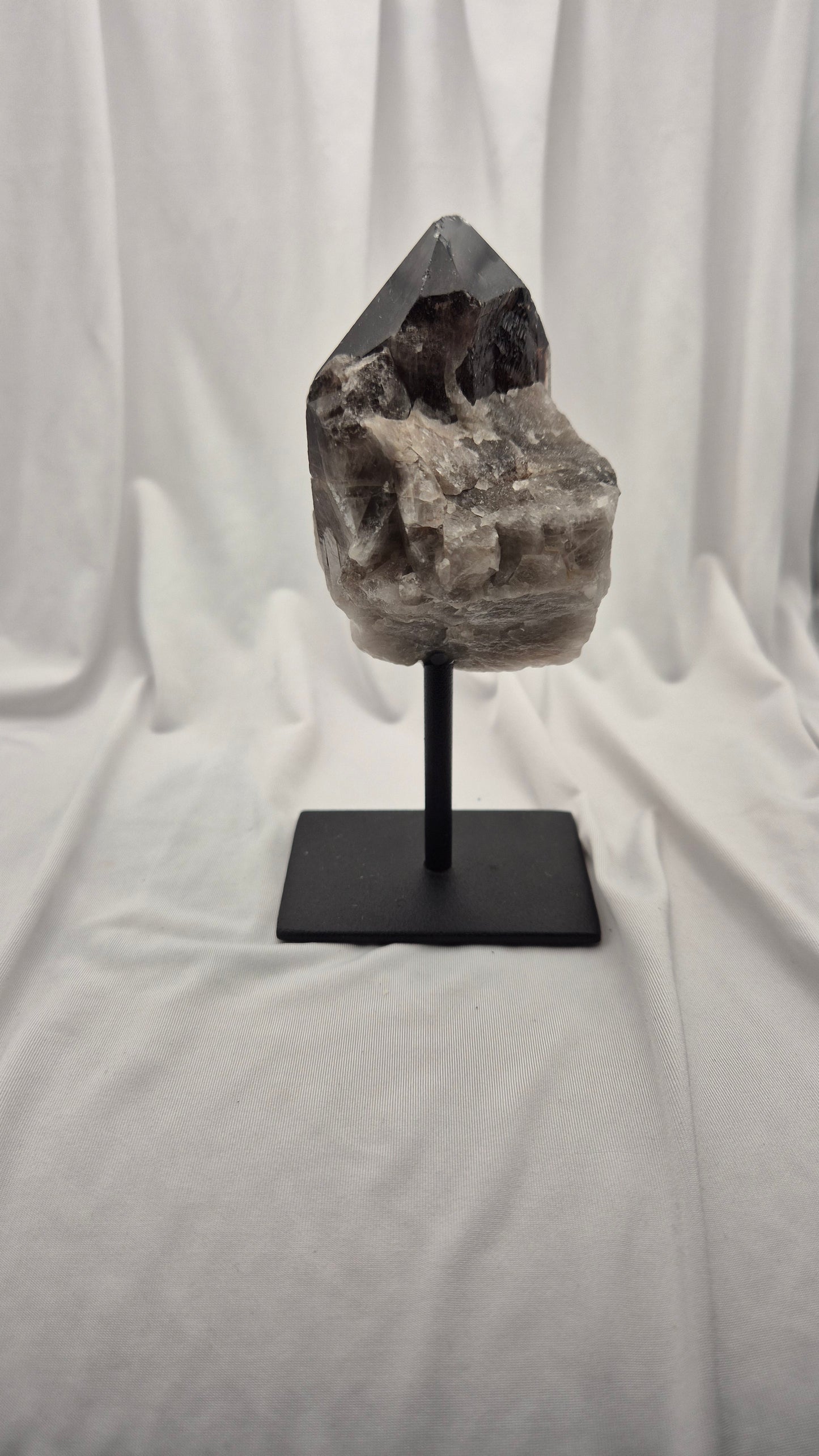 A natural smoky quartz crystal cluster on a black metal display stand, set against a draped white fabric backdrop.