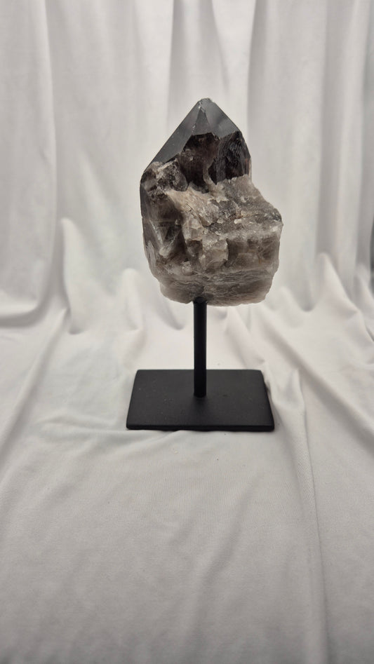 A natural smoky quartz crystal cluster on a black metal display stand, set against a draped white fabric backdrop.